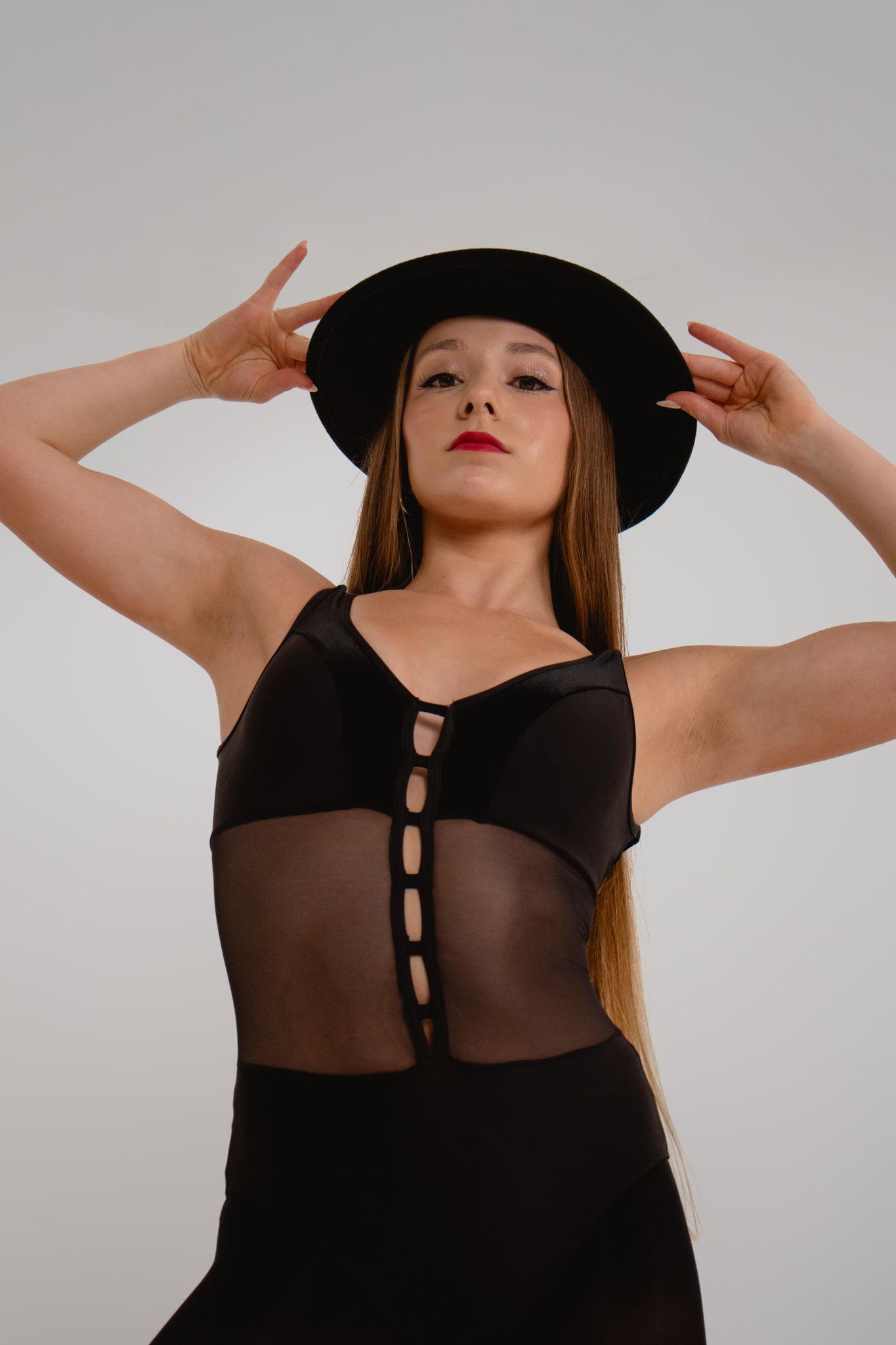 Dancer in black mesh bodysuit and wide-brim hat poses with hands framing face.