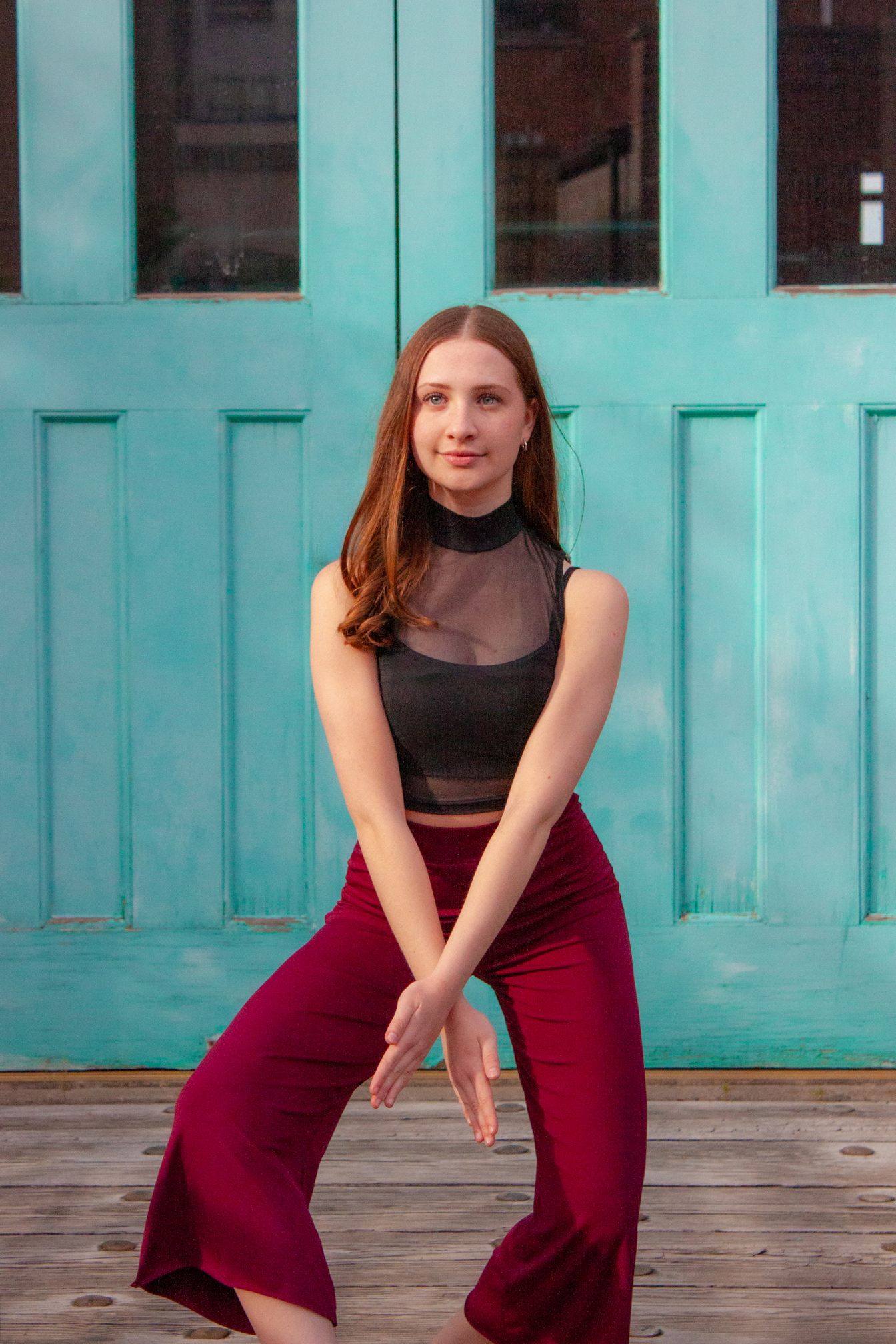 A young woman in a black mesh top and red flare pants poses gracefully in front of a teal door.