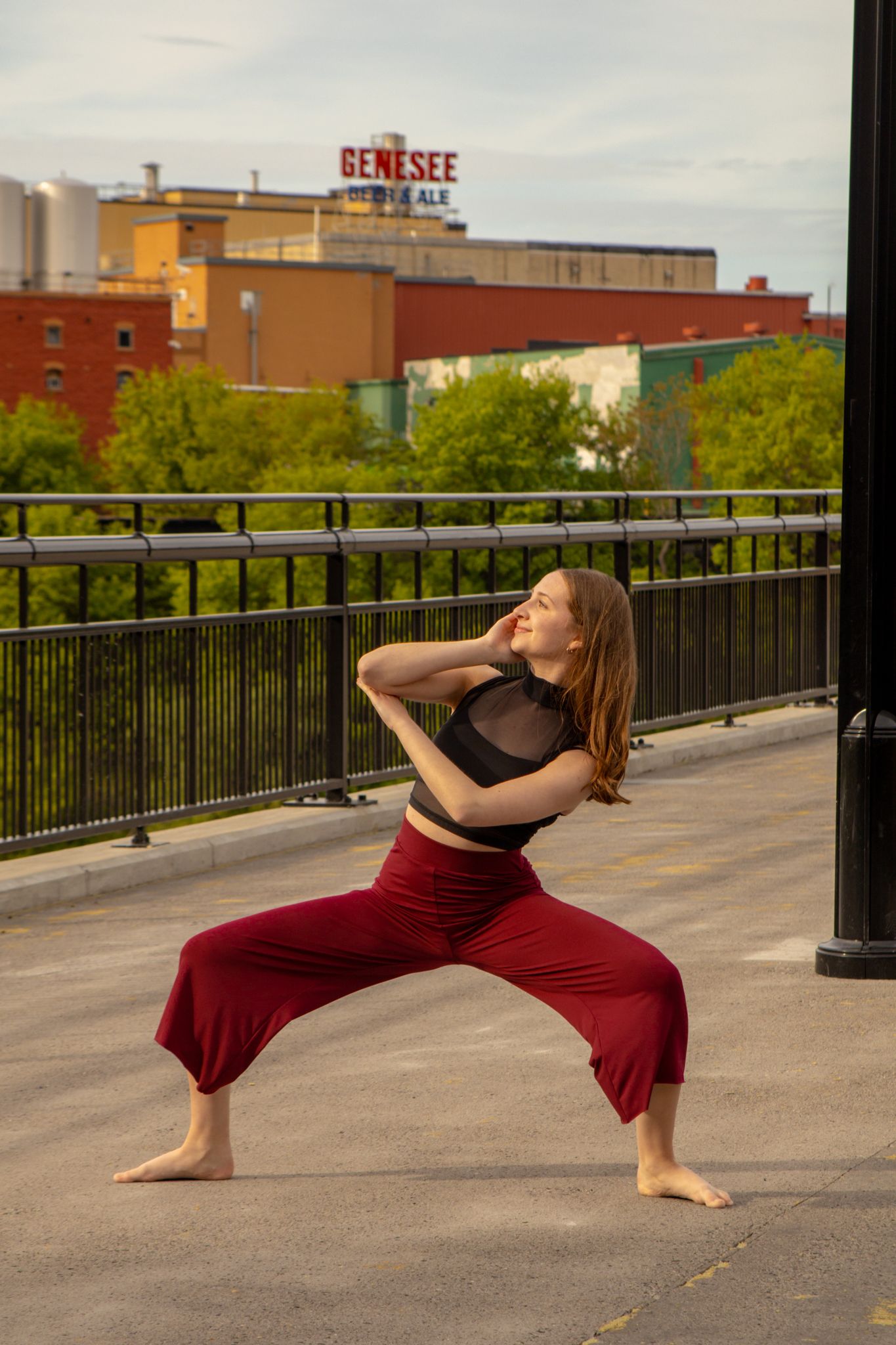 A barefoot dancer lunges in red wide-leg pants outdoors with the Genesee Brewery building visible in the background.