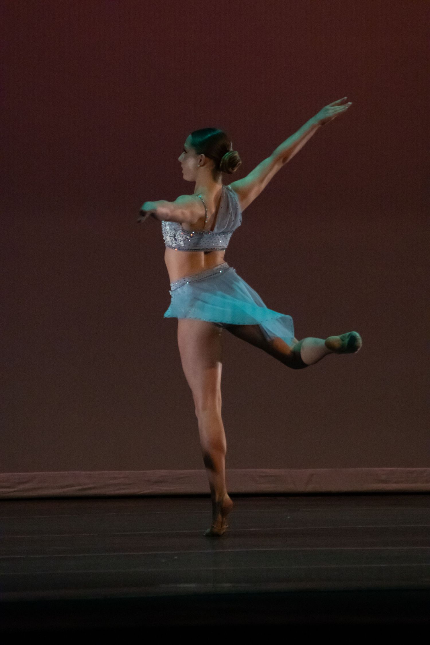 A solo dancer in a turquoise skirt and silver top performs an elegant arabesque on stage.