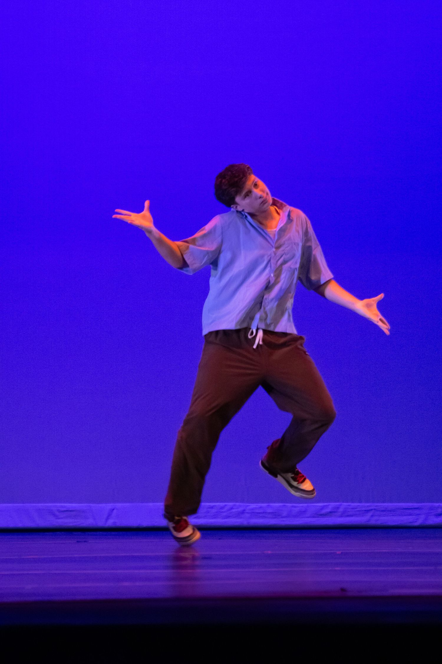 A male hip-hop dancer balances on one foot with arms outstretched on a blue-lit performance stage.