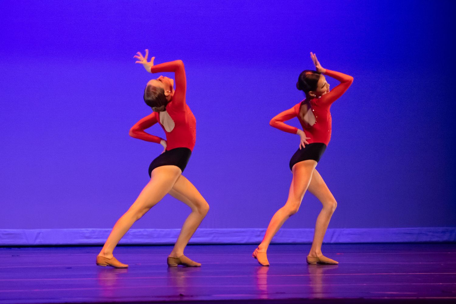 Two dancers in matching red costumes perform a synchronized contemporary routine on a blue-lit stage.