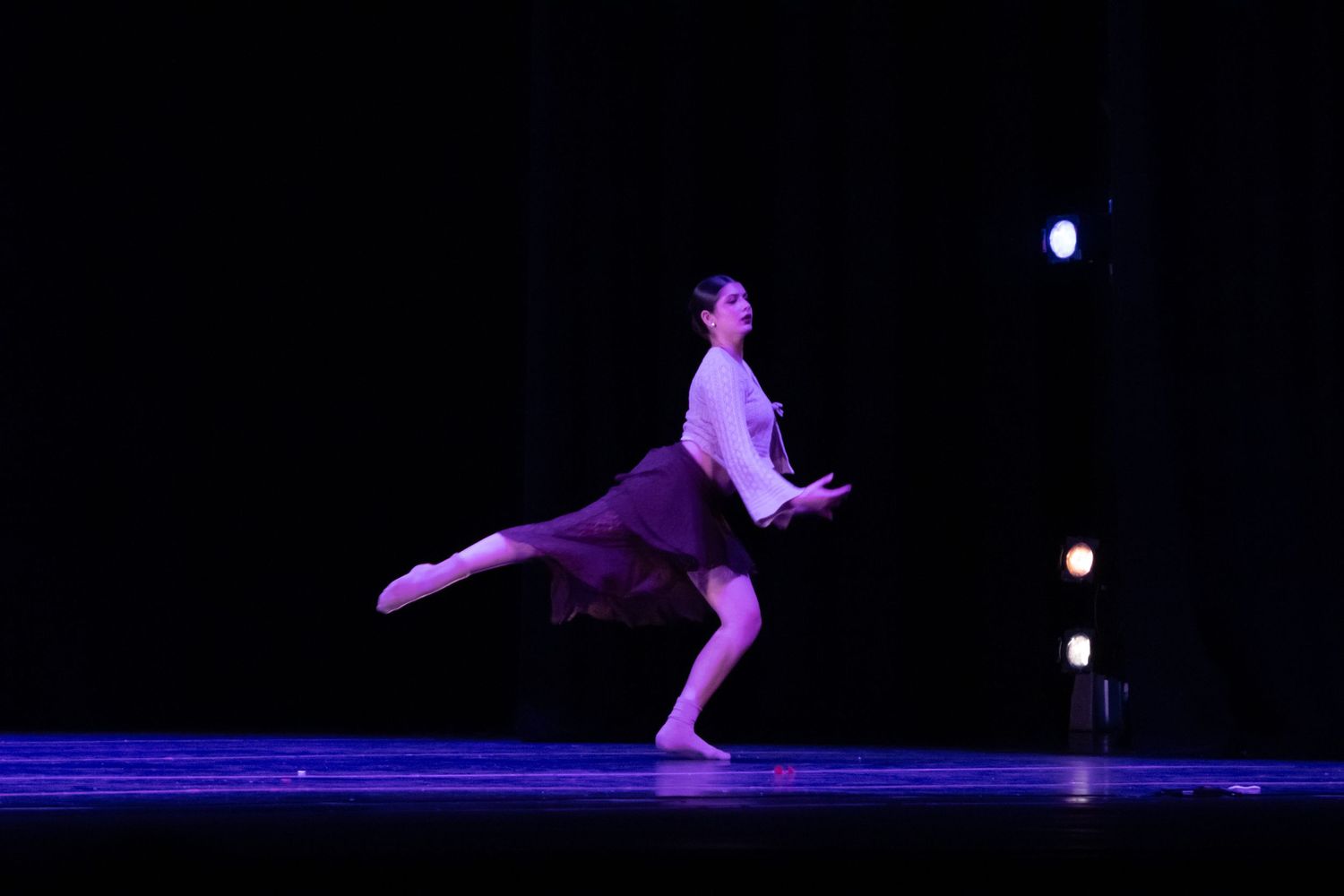 Female dancer in white top and brown skirt performs solo on dimly lit blue stage.