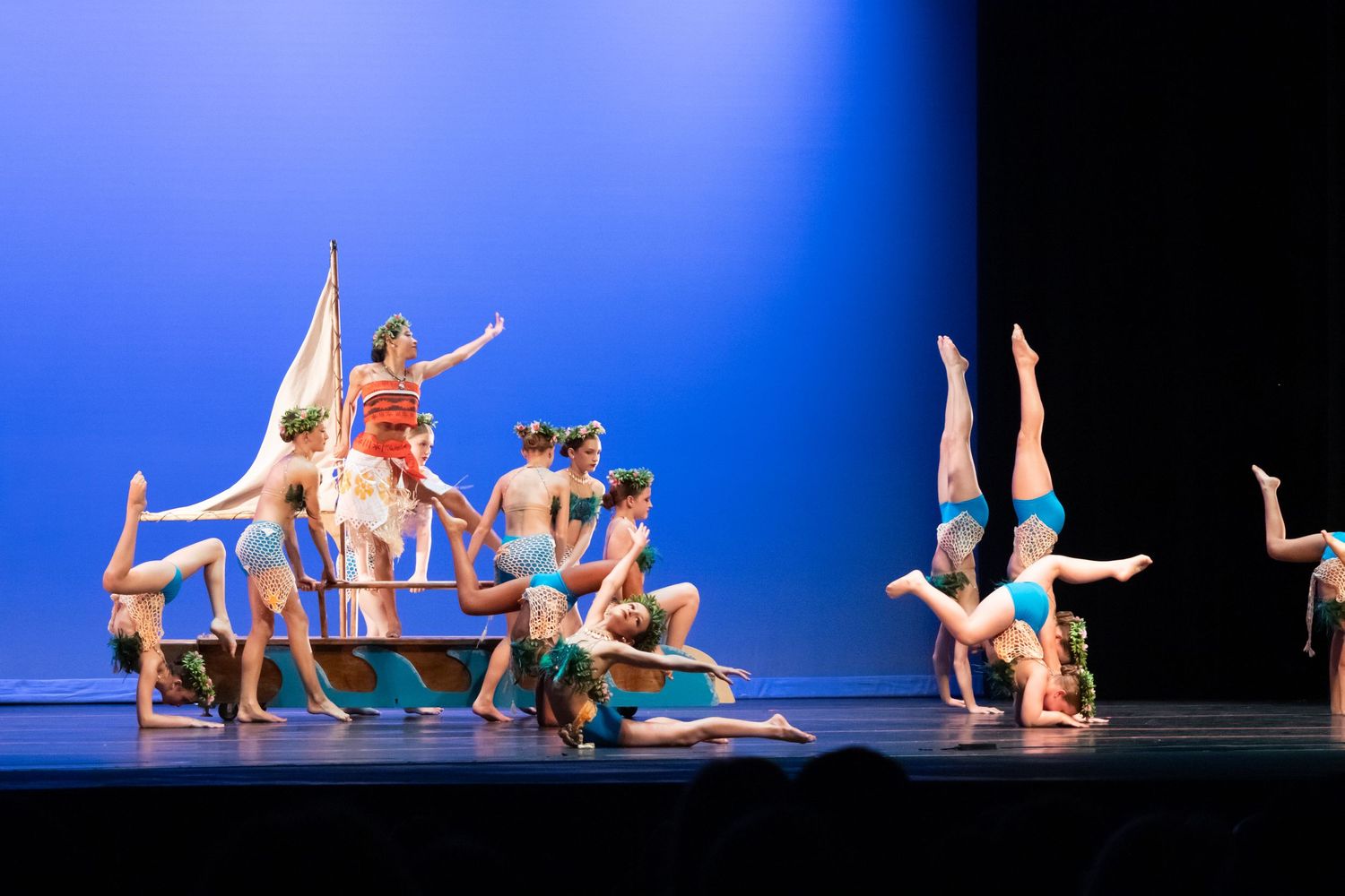 A group of dancers in colorful costumes perform acrobatic moves on a bright blue-lit stage.