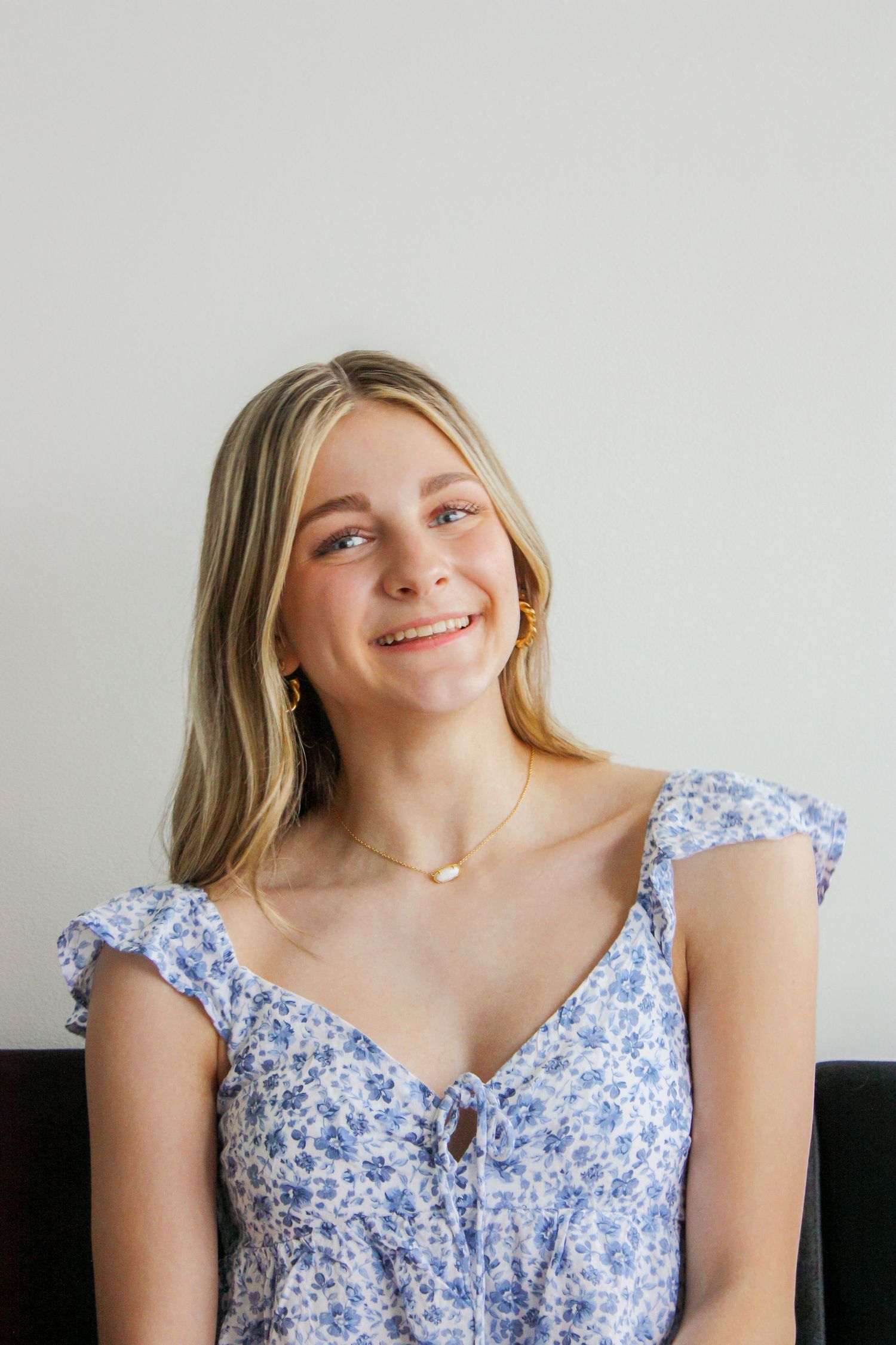 Smiling young blonde woman wearing a blue floral dress and gold necklace sitting against a white background.