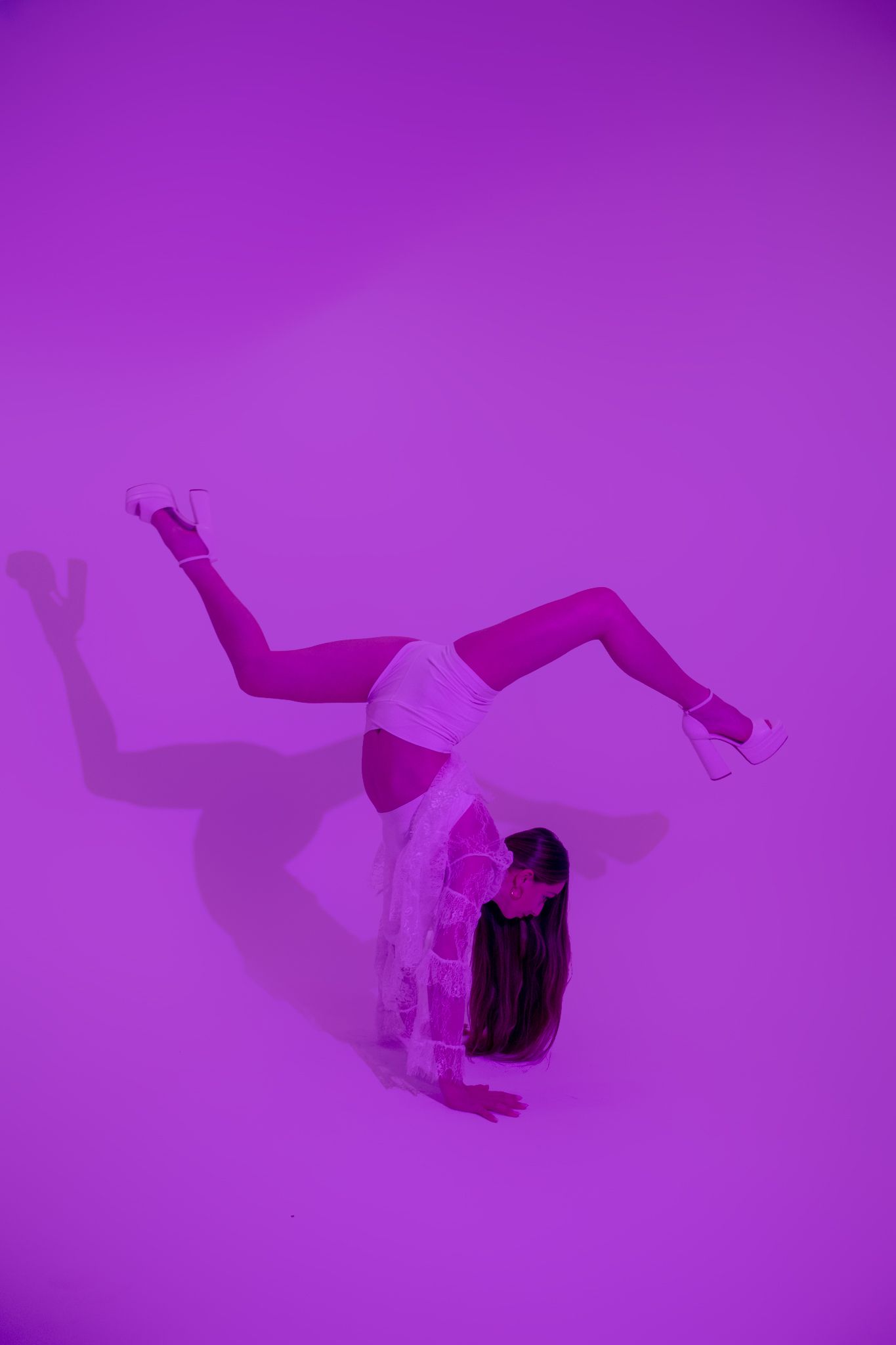 A dancer strikes an acrobatic pose with legs raised against a vivid purple background wearing a white outfit and heels.