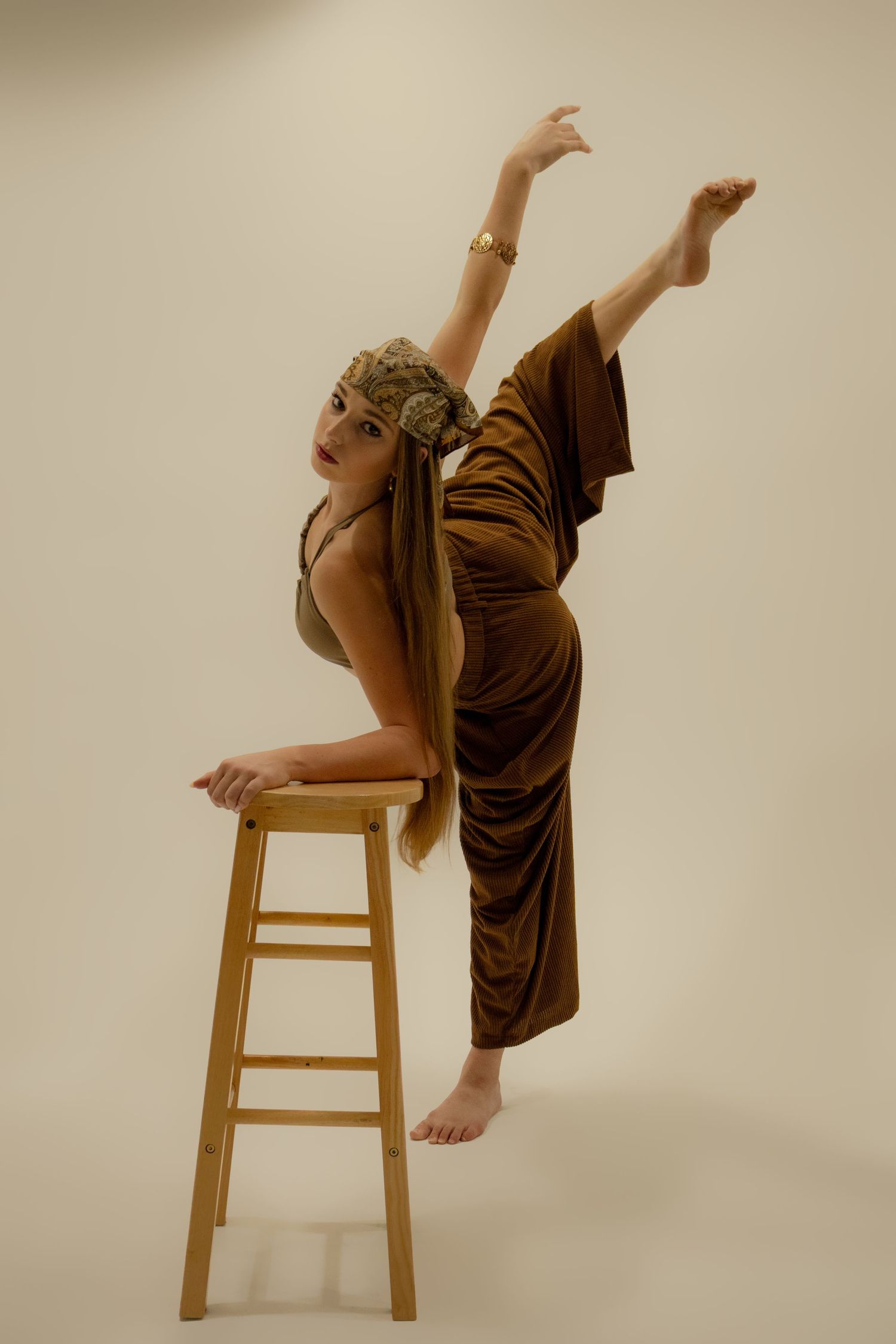 Flexible dancer in brown outfit balances leg vertically while leaning on wooden stool in studio.