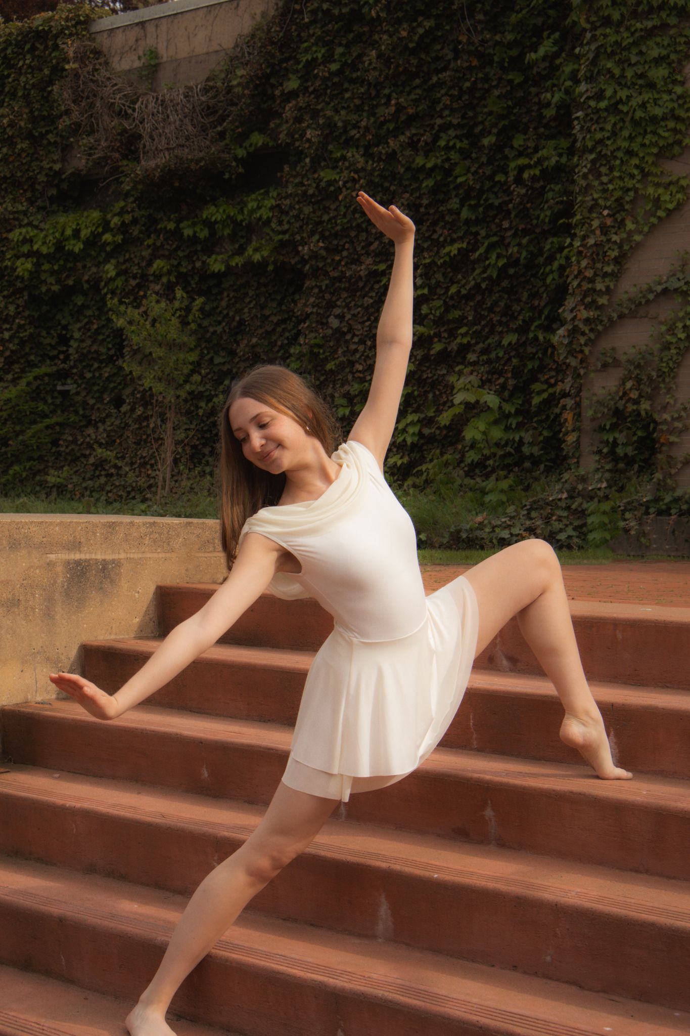 Barefoot dancer in white dress performs elegant side stretch on outdoor brick staircase with ivy.