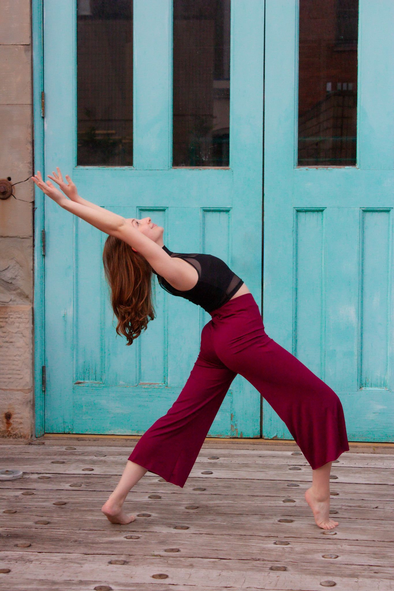 Female contemporary dancer arches backward gracefully in front of a teal door wearing burgundy wide-leg pants.