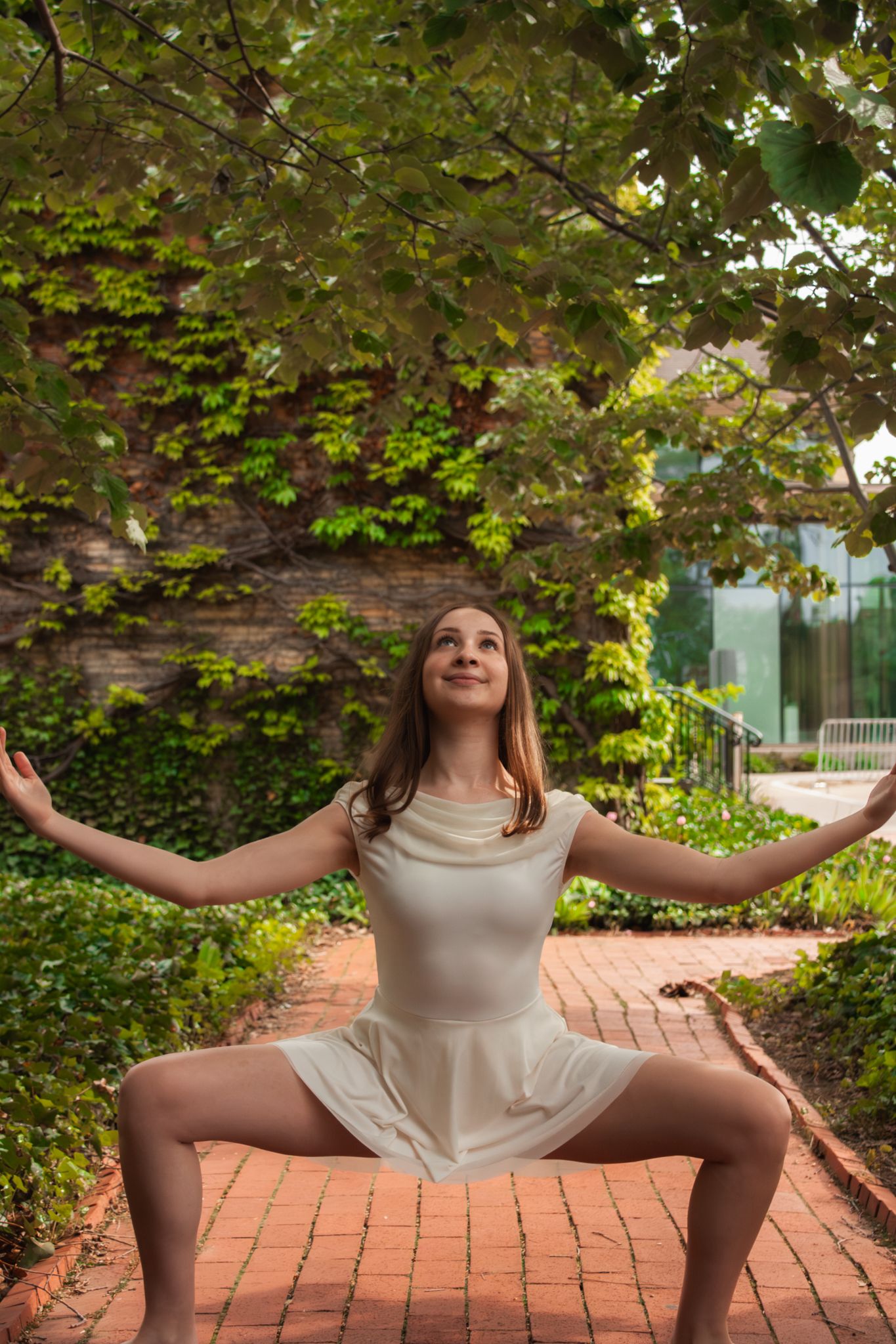 Young dancer in white dress performing wide plie pose on brick pathway under green leafy tree canopy.