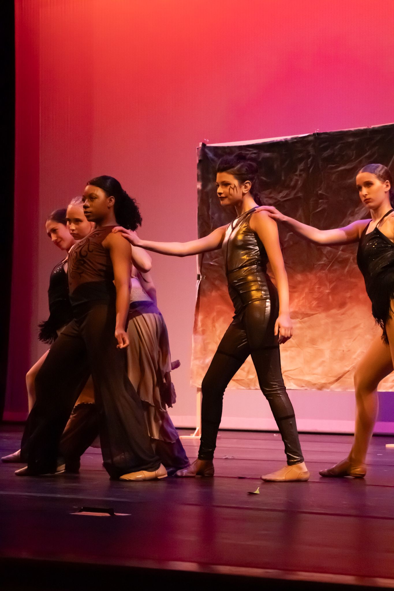Contemporary dance performers in black and gold costumes move across a stage lit with warm red lighting.