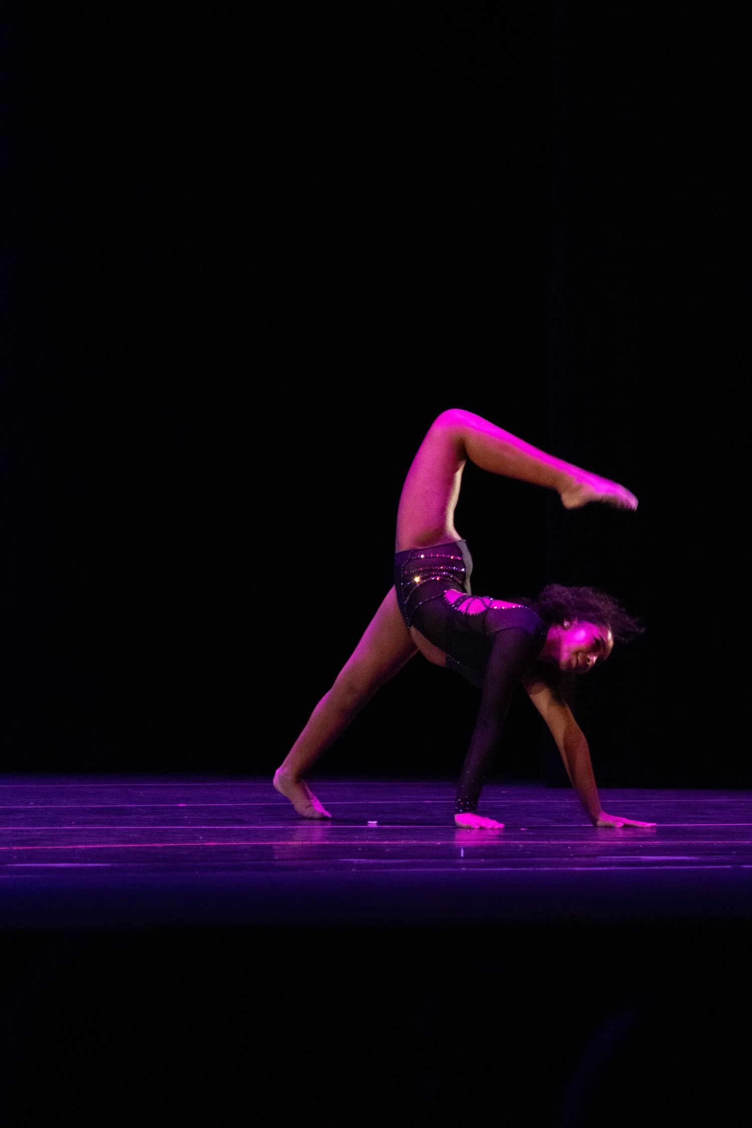 A flexible dancer performs an acrobatic move under dramatic pink stage lighting on a dark stage.