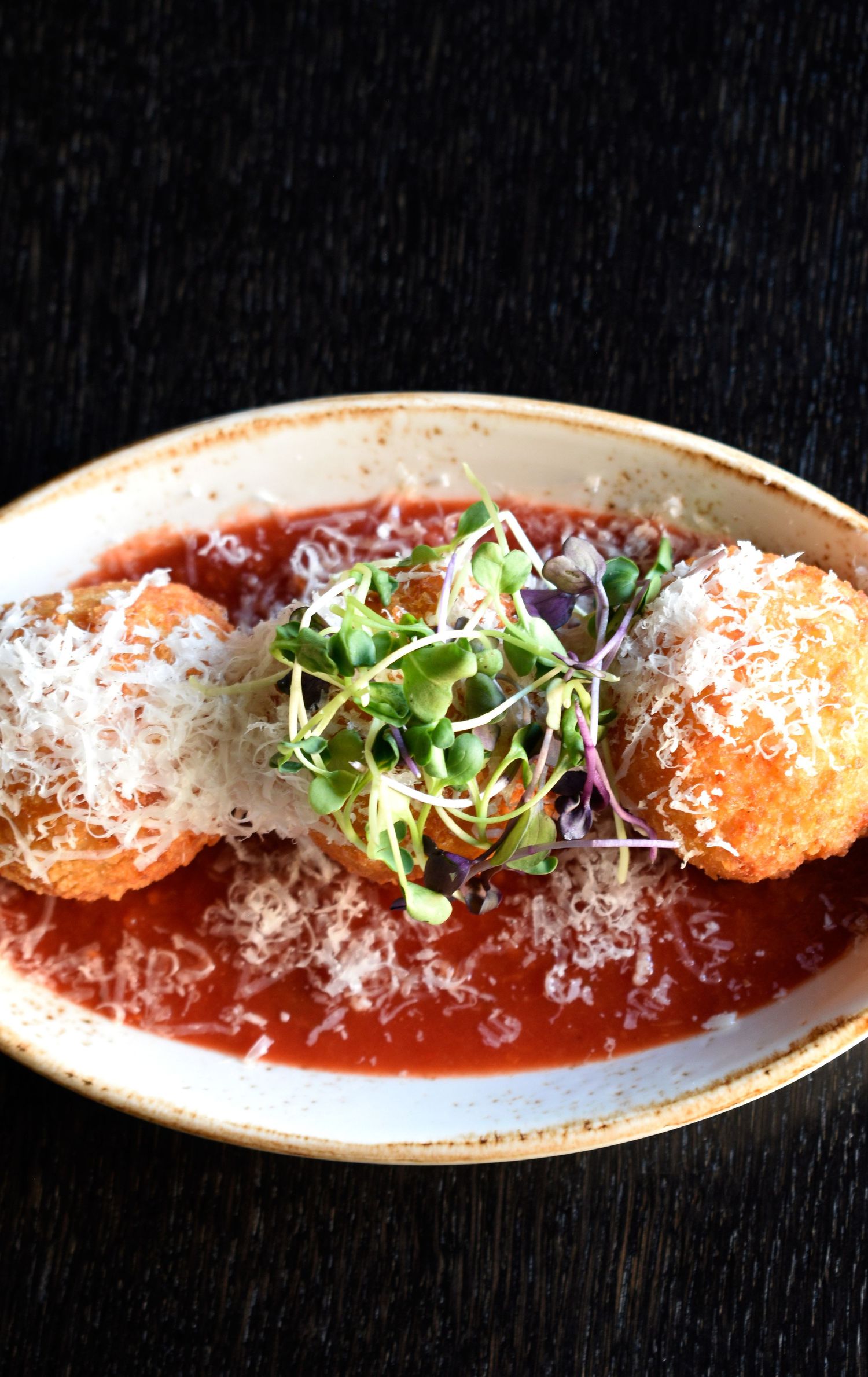Golden fried arancini in tomato sauce topped with grated parmesan and fresh microgreens on a plate.