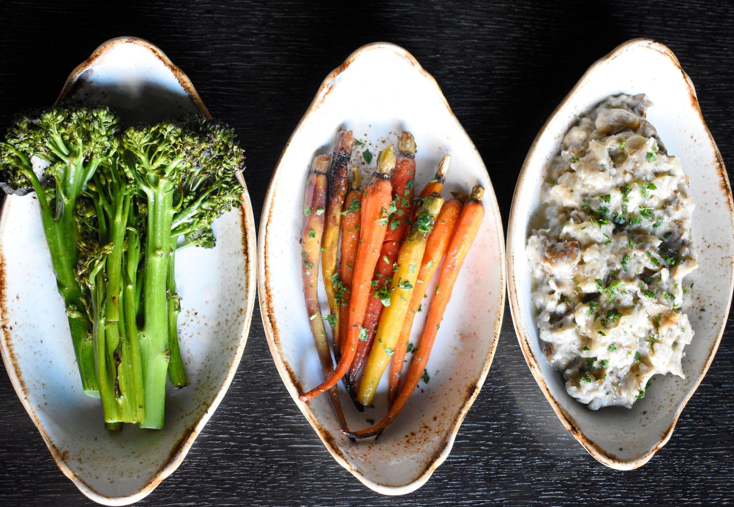 Three oval dishes with roasted broccolini, glazed carrots, and creamy herb side dishes on dark surface.