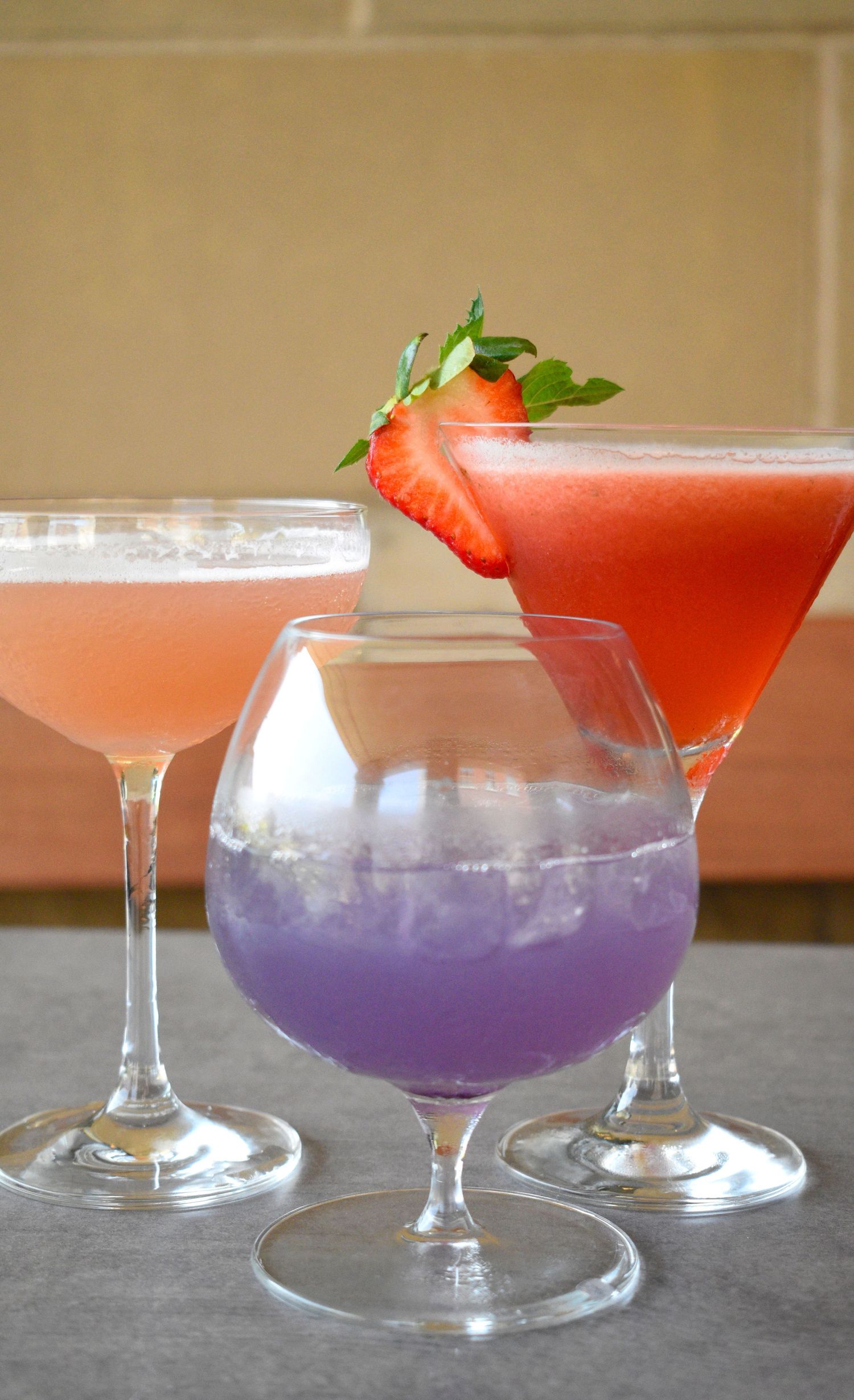 Three vibrant cocktails in pink, purple, and red hues garnished with strawberry and mint on a bar.