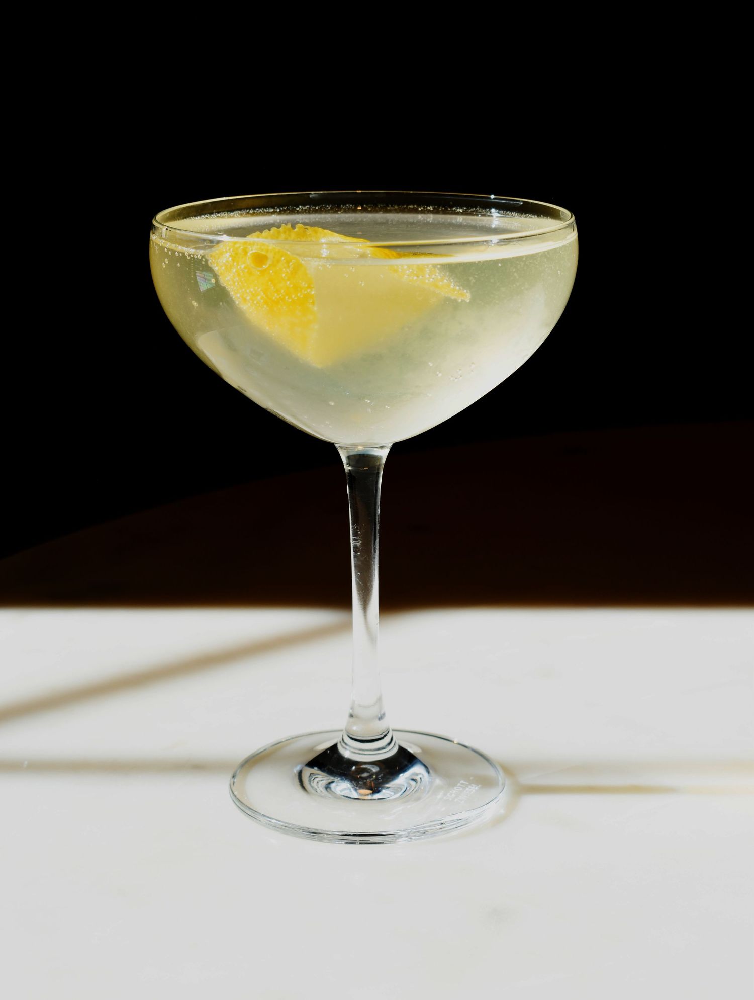 An elegant coupe glass containing a pale green cocktail garnished with a twisted lemon peel against a dark background.