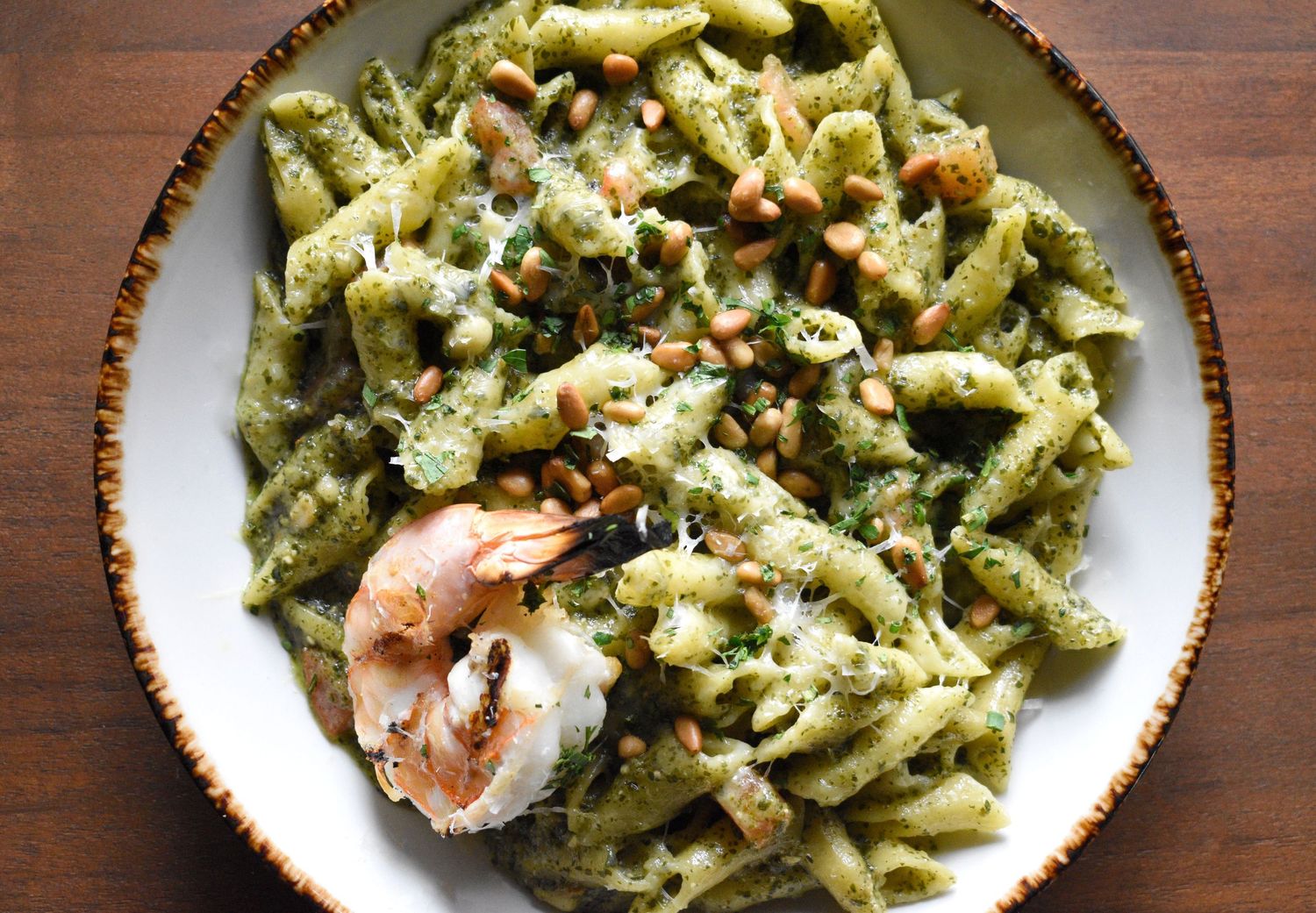 Penne pasta with pesto sauce, grilled shrimp, pine nuts, and parmesan cheese in a rustic bowl.