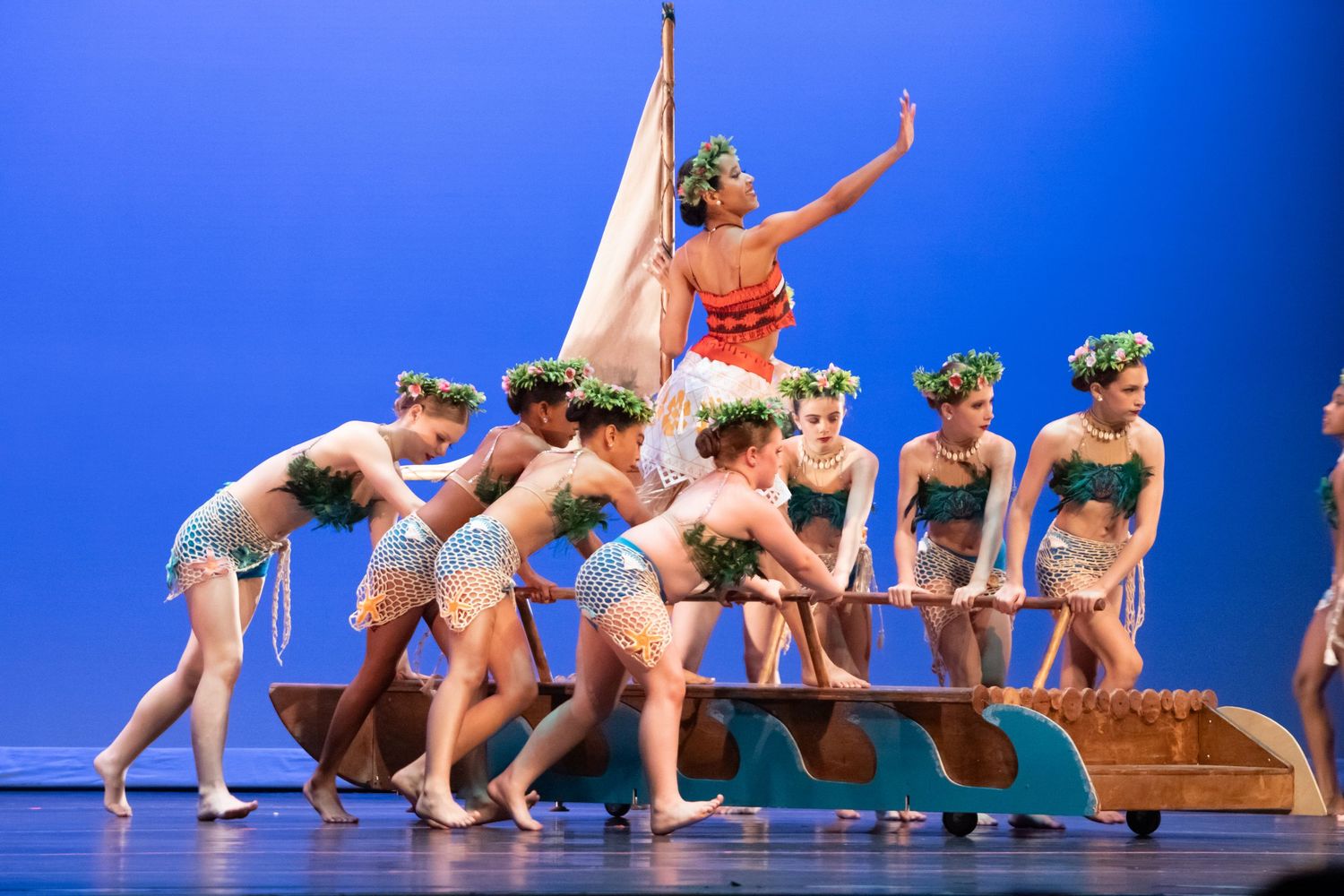 A group of dancers in tropical costumes perform a Moana-inspired routine with a boat prop on a blue-lit stage.