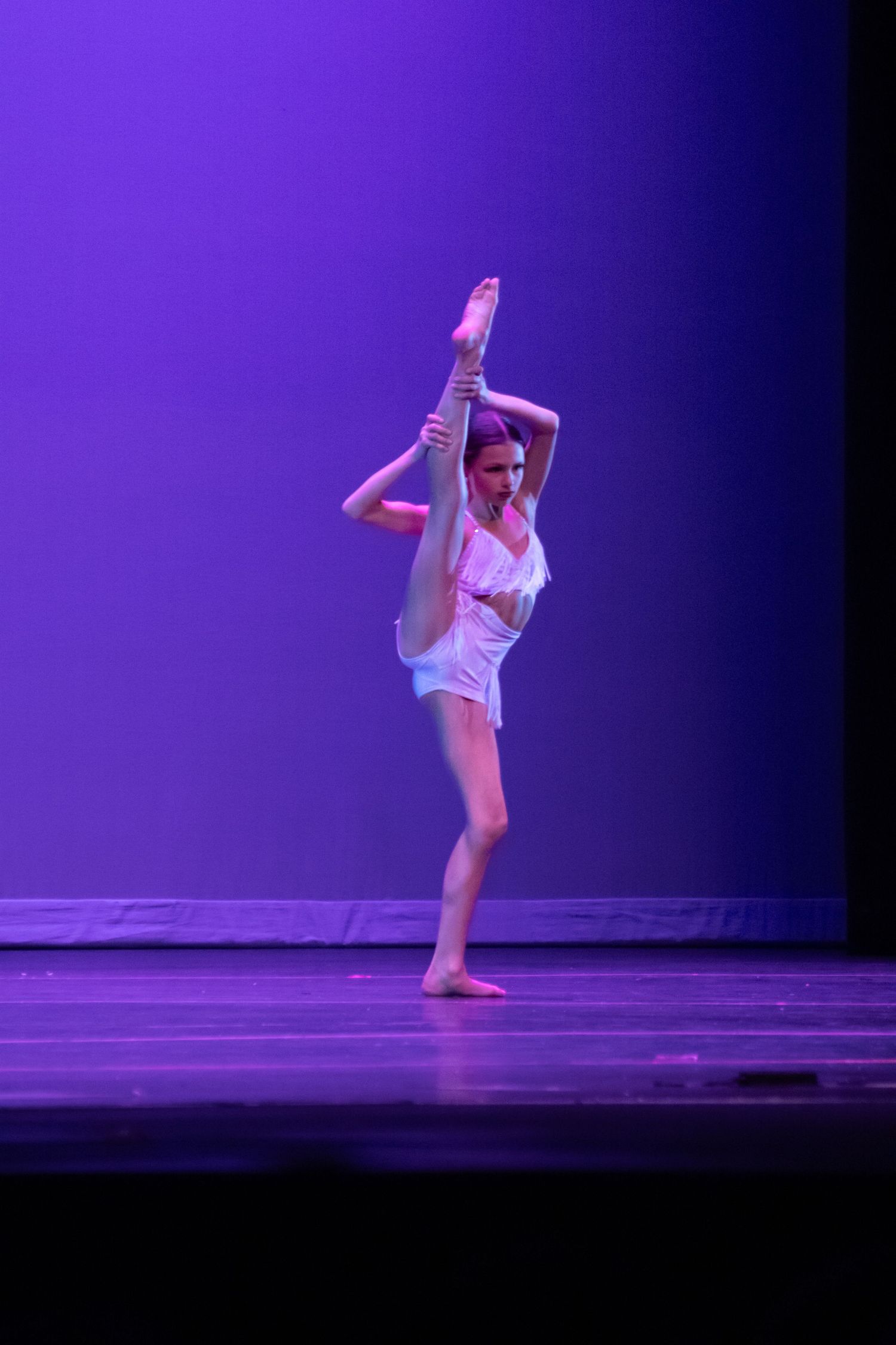 A flexible dancer holds a leg extension pose on a purple-lit stage in a white outfit.