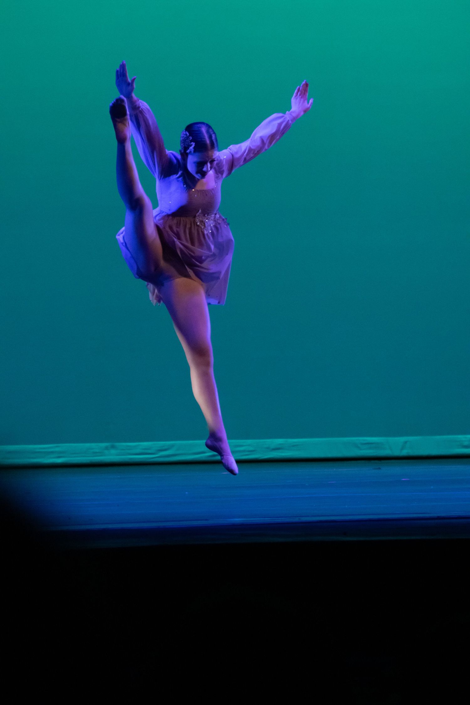 Dancer in purple costume executes high leg extension jump under teal stage lighting.