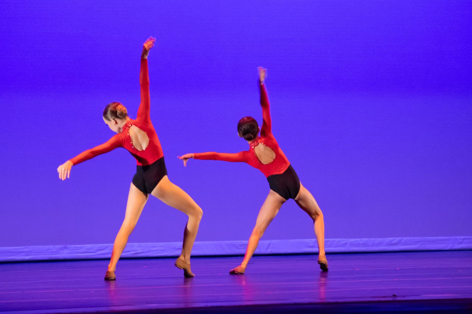 Two female dancers in red backless leotards perform jazz dance moves on a blue-lit stage.
