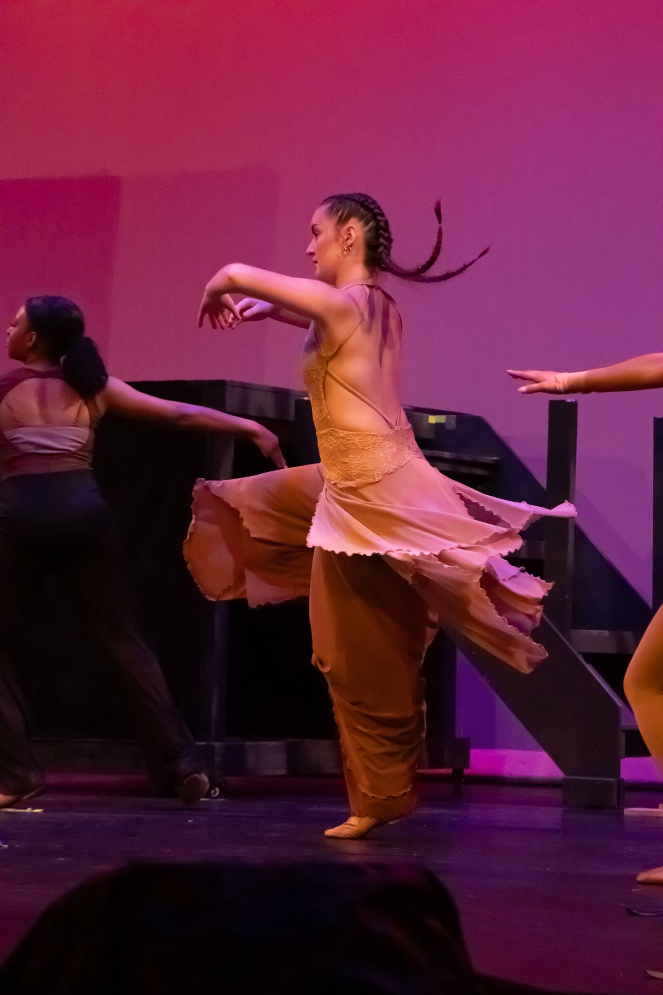 A dancer in a flowing peach dress performs an expressive contemporary dance move on a pink-lit stage.