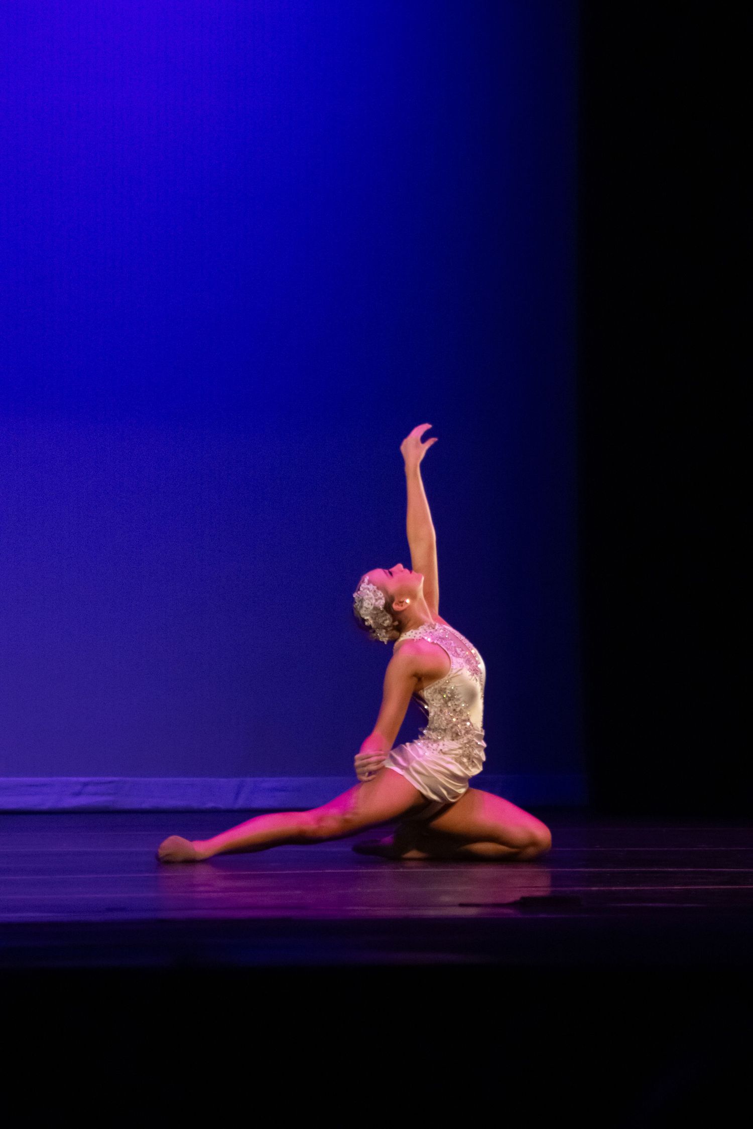 A dancer in a white costume performs an elegant floor pose with arm raised under blue stage lighting.