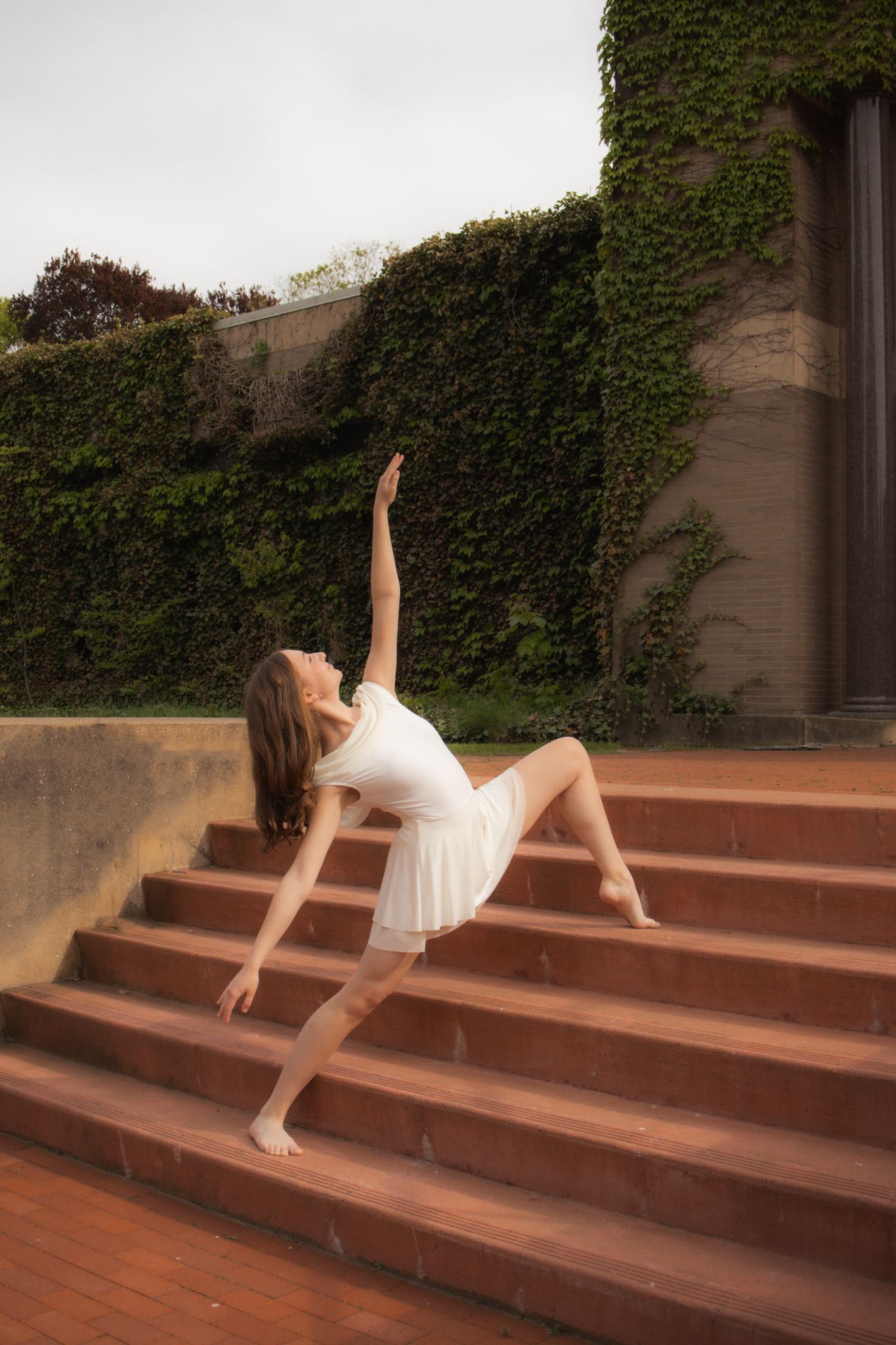 Ballerina in white dress performing elegant pose on brick steps with ivy-covered wall.