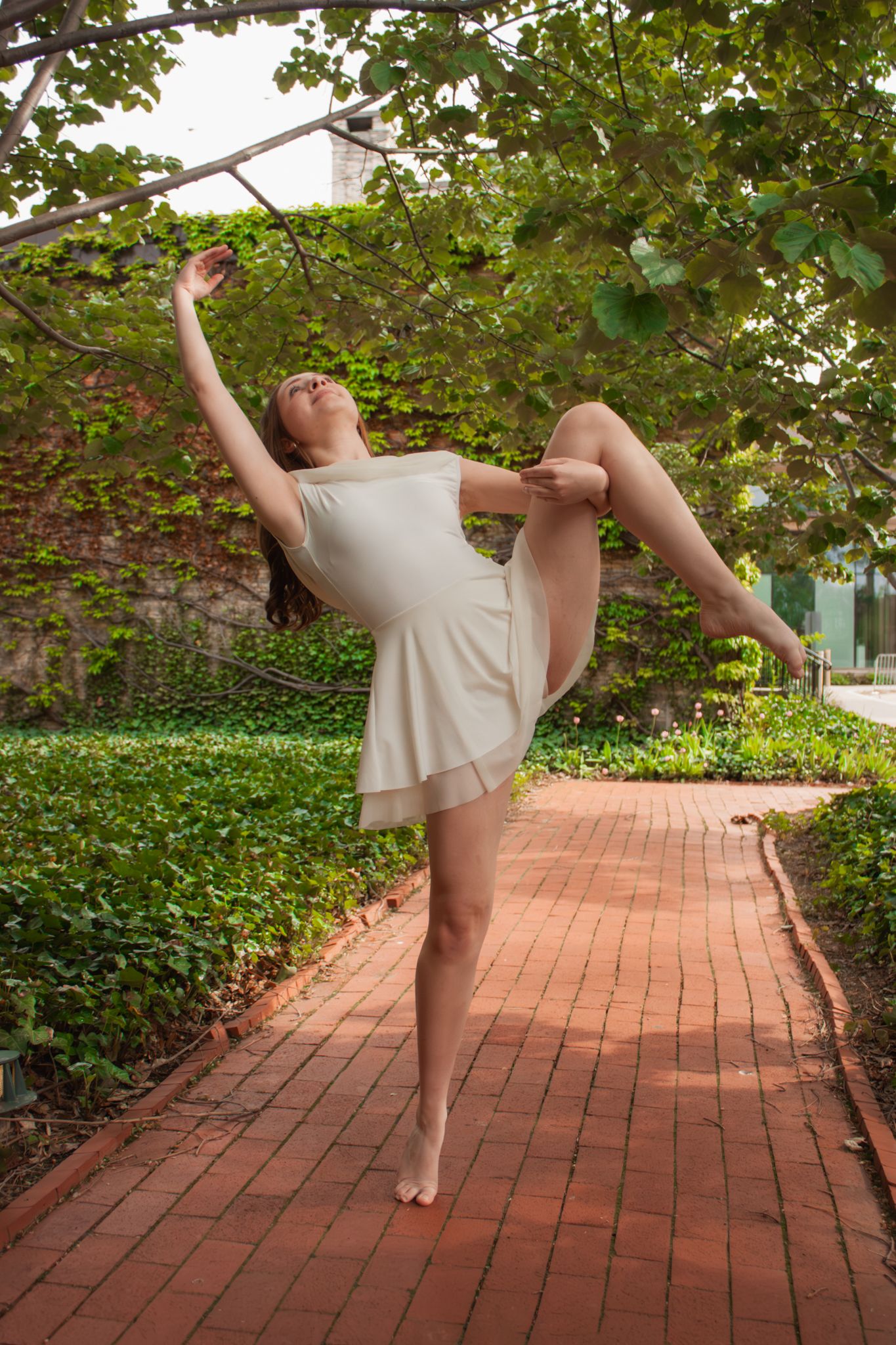 A graceful dancer performs a high leg extension in a white dress on a brick path surrounded by lush green foliage.