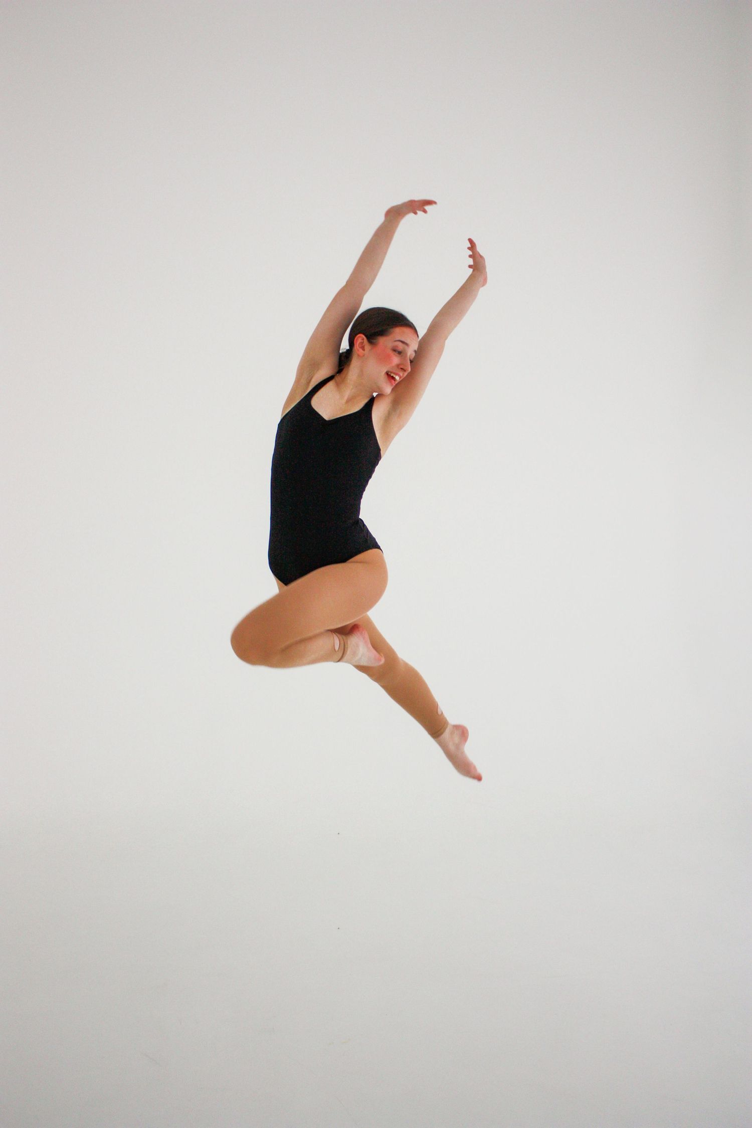 A female dancer in a black leotard performs a dynamic mid-air jump with arms raised on a white background.