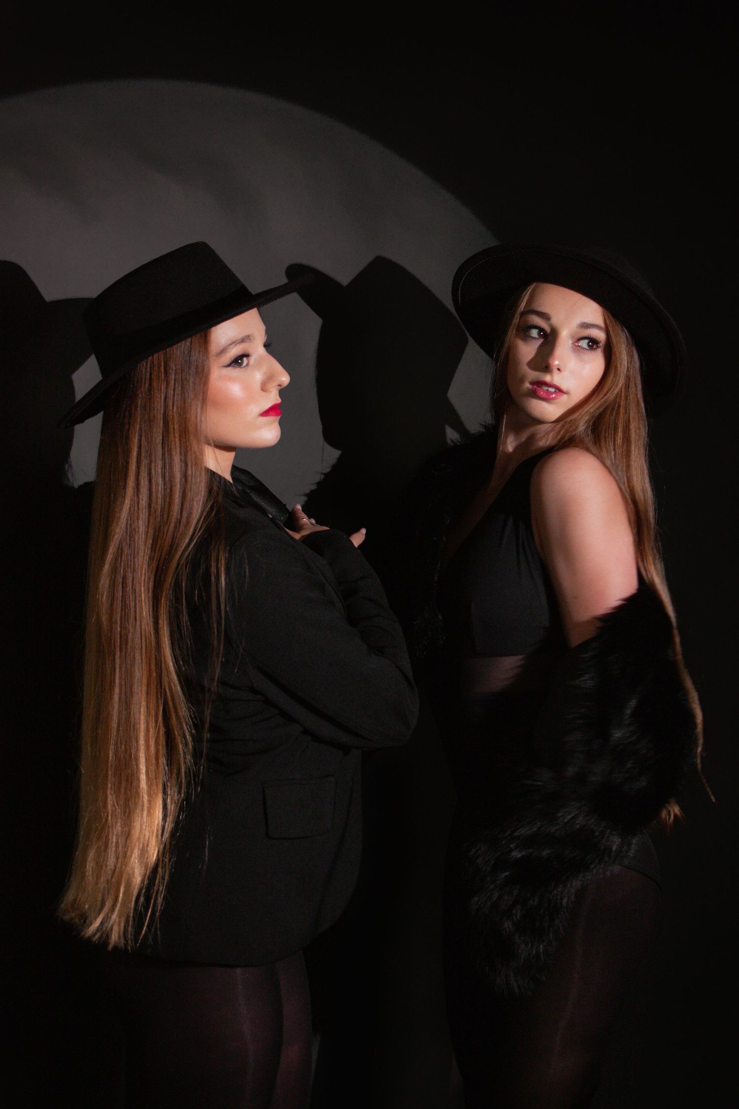Two stylish women in black outfits and wide-brim hats pose back-to-back against a dark dramatic background.