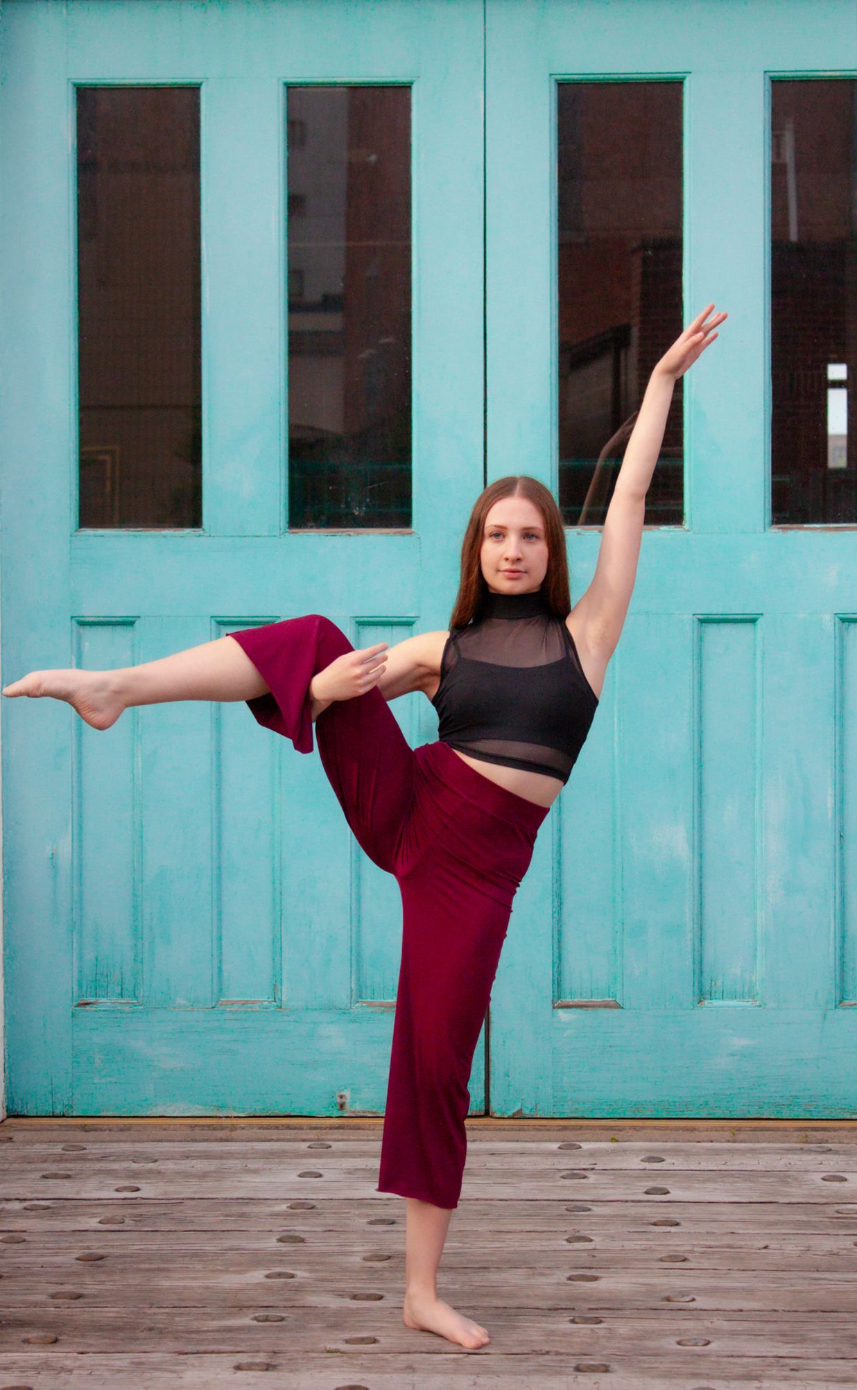 Young female dancer balancing on one leg in a high kick pose, wearing burgundy pants and black top before teal doors.