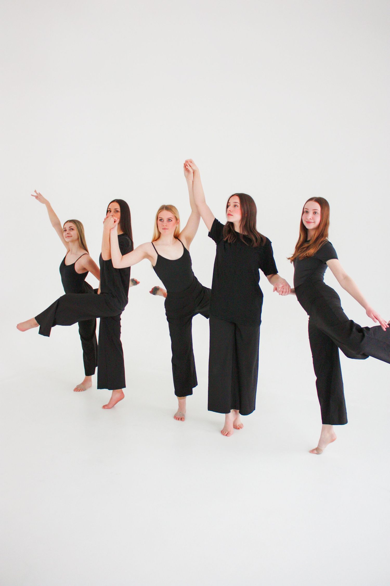 Five female dancers in black outfits perform a synchronized leg lift routine against a white background.
