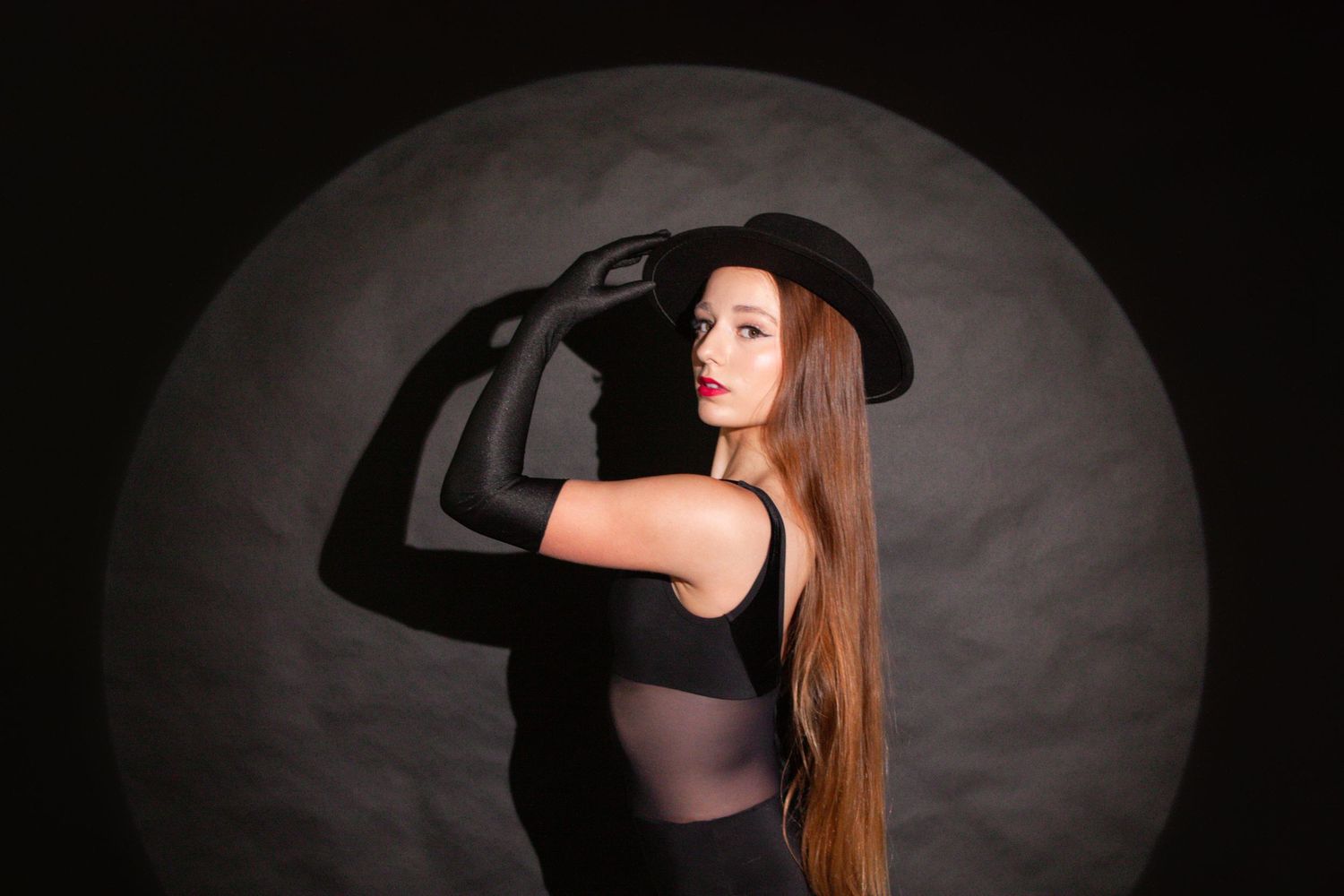 Woman in black bodysuit and wide-brim hat poses dramatically against dark circular spotlight background.