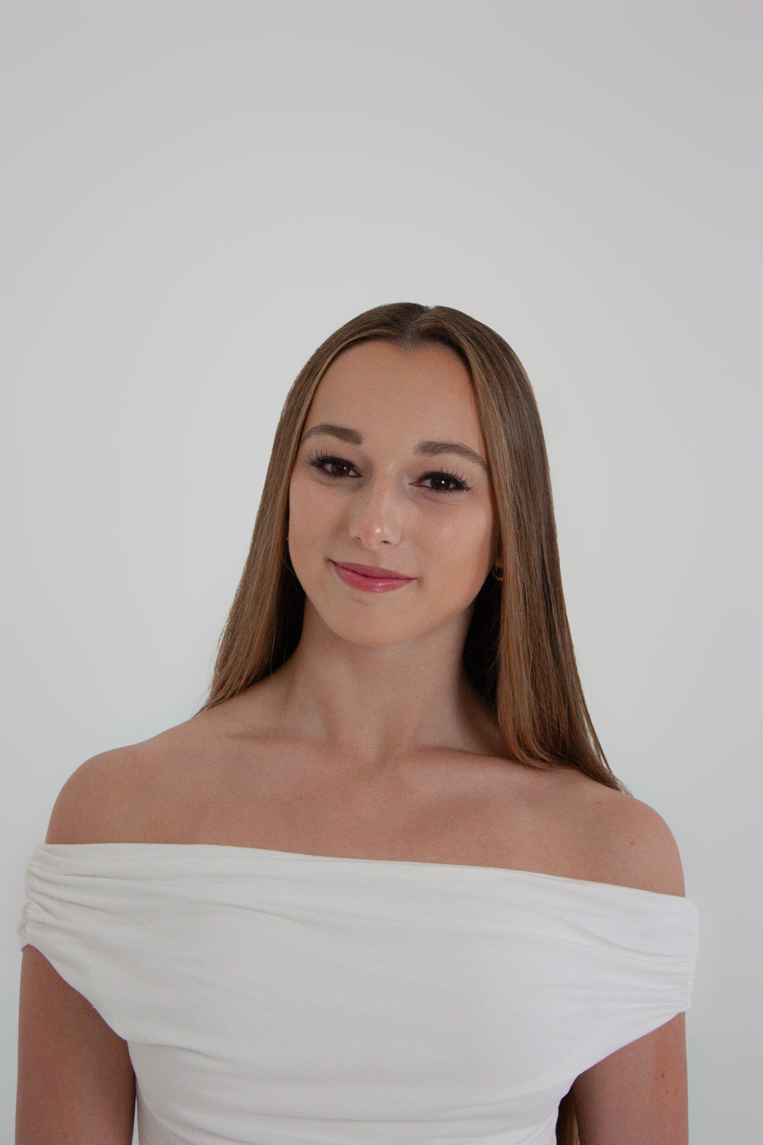 Young woman with long straight brown hair wearing a white off-shoulder dress poses against a light gray background.