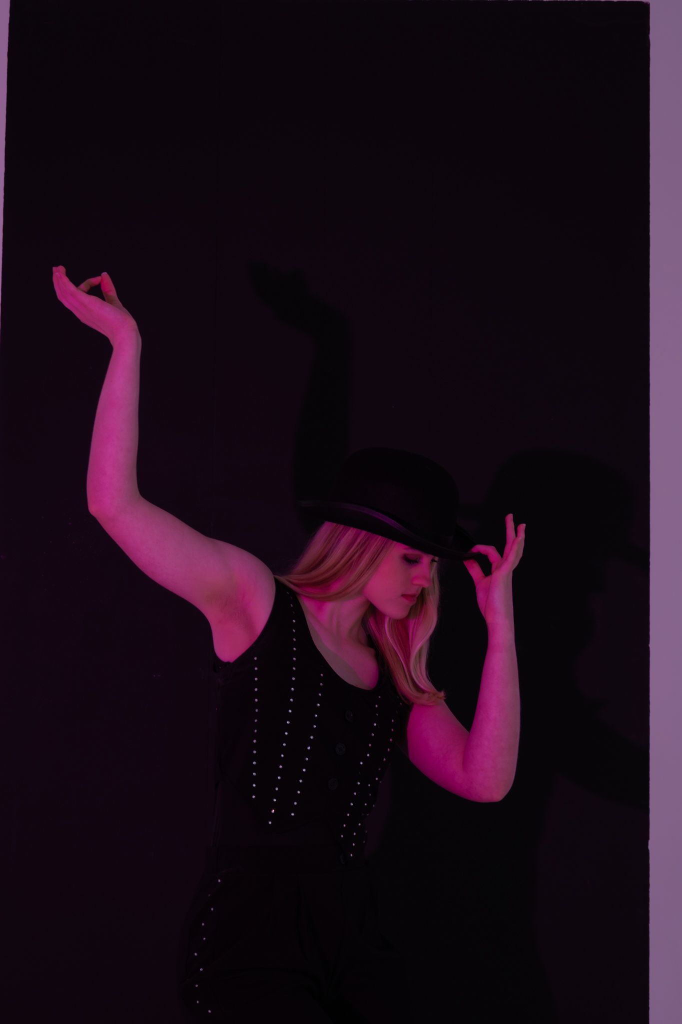 Blonde dancer in black studded outfit and hat poses dramatically under pink-purple studio lighting.