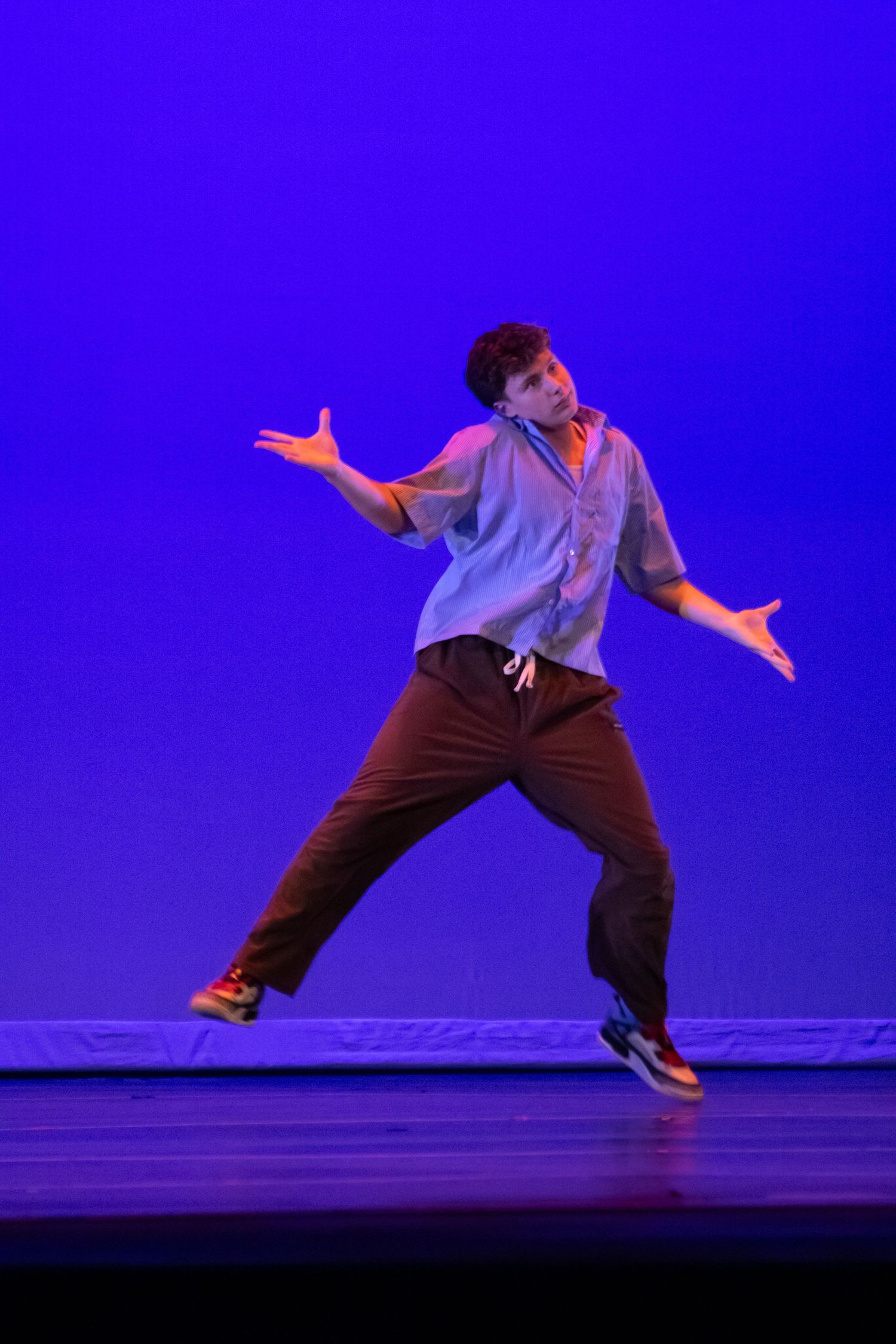 A male dancer performs hip-hop choreography in casual attire on a blue-lit stage.
