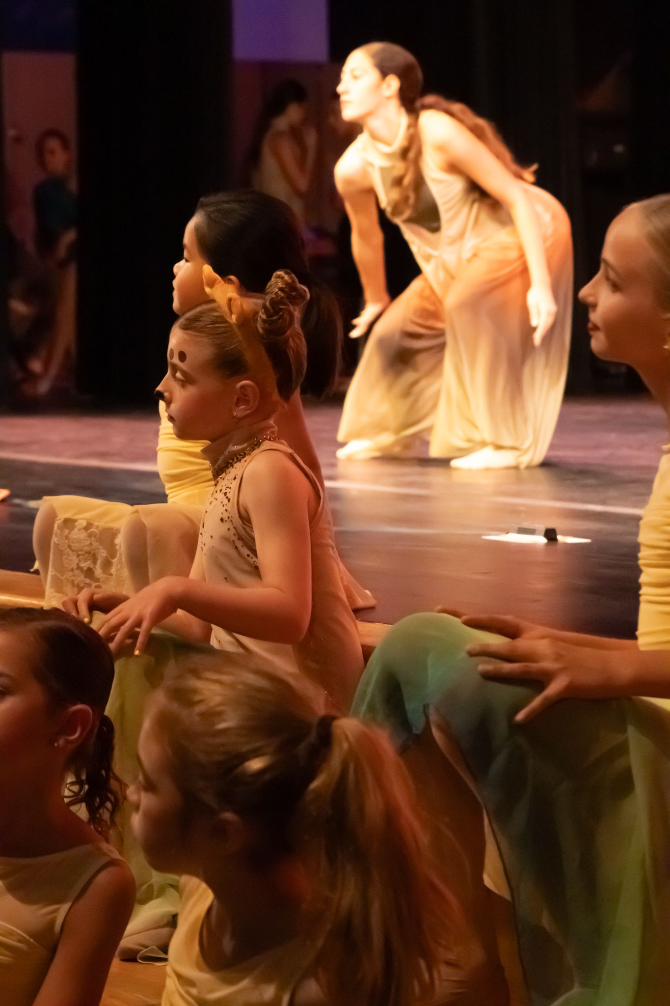 Young dancers in golden costumes perform on stage while older dancers crouch gracefully in the background.