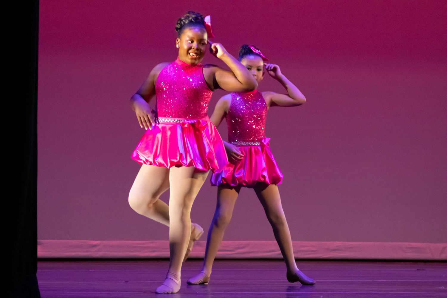 Two girls in pink sequined dresses perform a jazz dance routine on a theatrical stage.