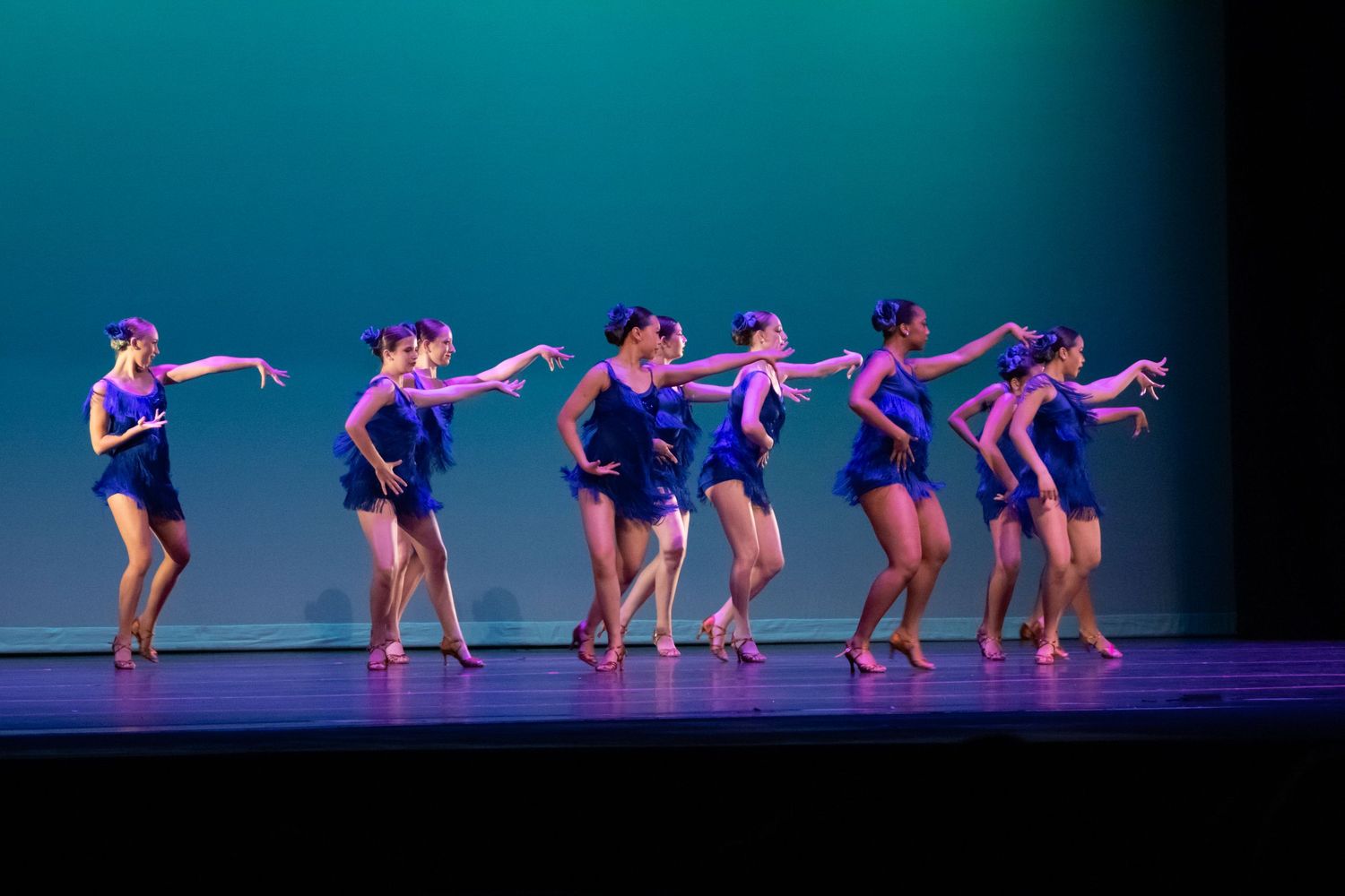 Seven female dancers in blue fringe costumes perform a synchronized routine on a teal-lit stage.