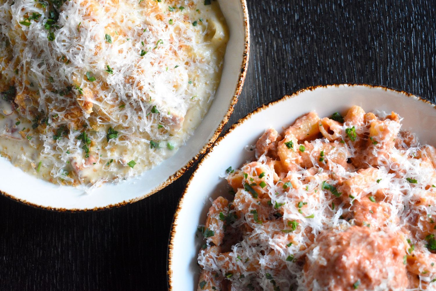 Two bowls of creamy pasta dishes topped with freshly grated Parmesan cheese on a dark table.