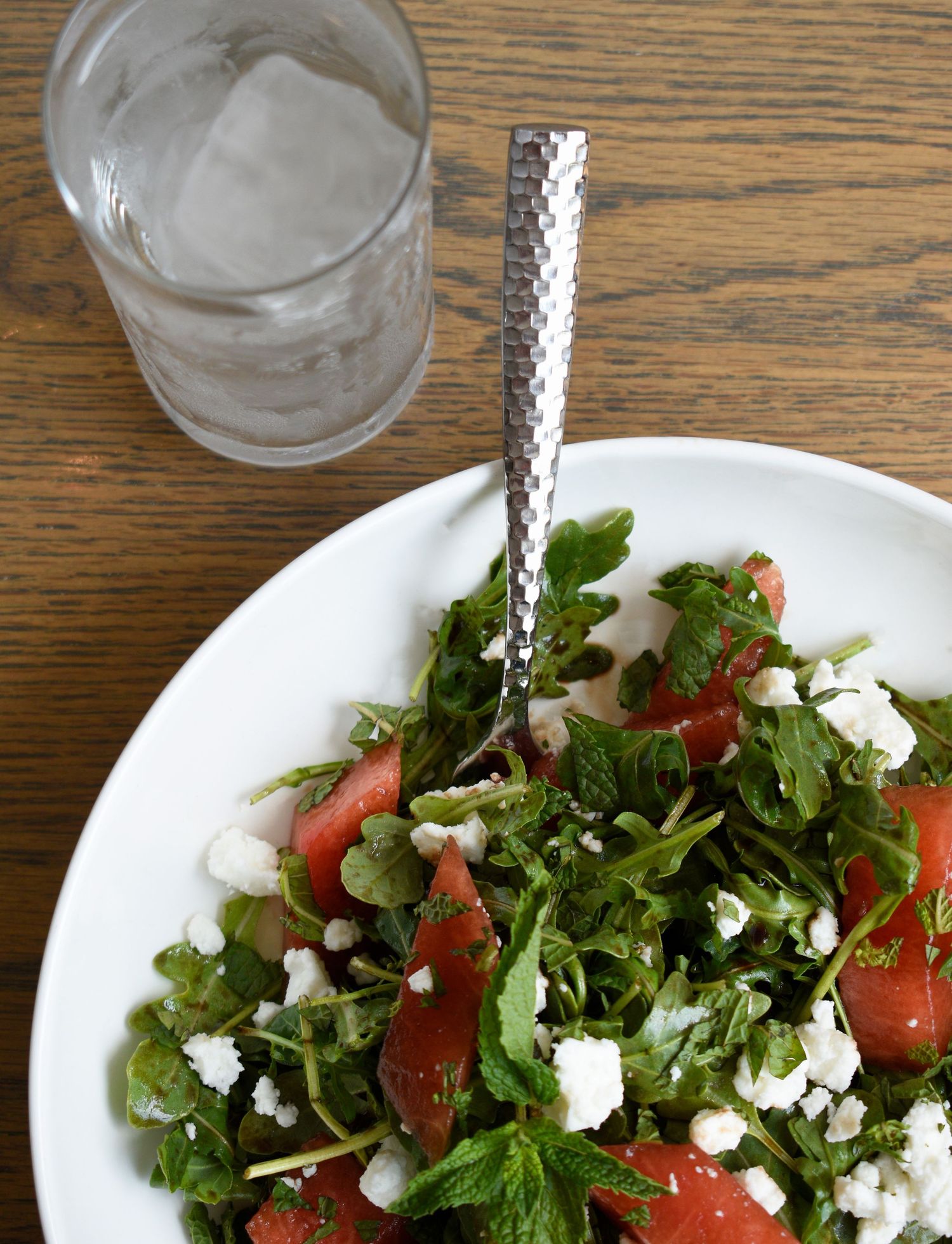Fresh watermelon salad with arugula and crumbled feta cheese served in a white bowl with water glass.