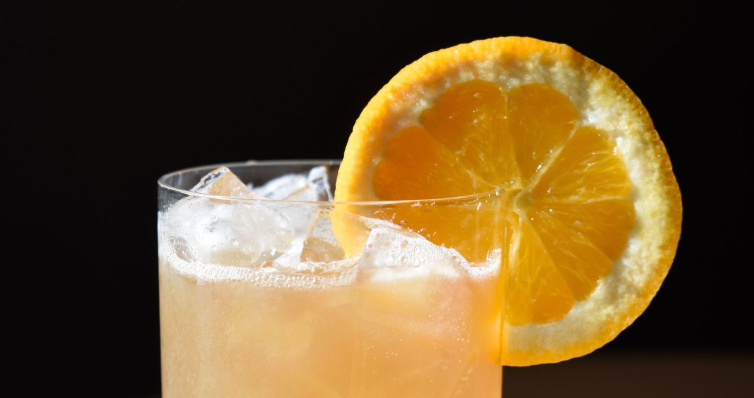 A frosty rocks glass with a pale orange cocktail on ice, garnished with a vibrant orange citrus wheel.
