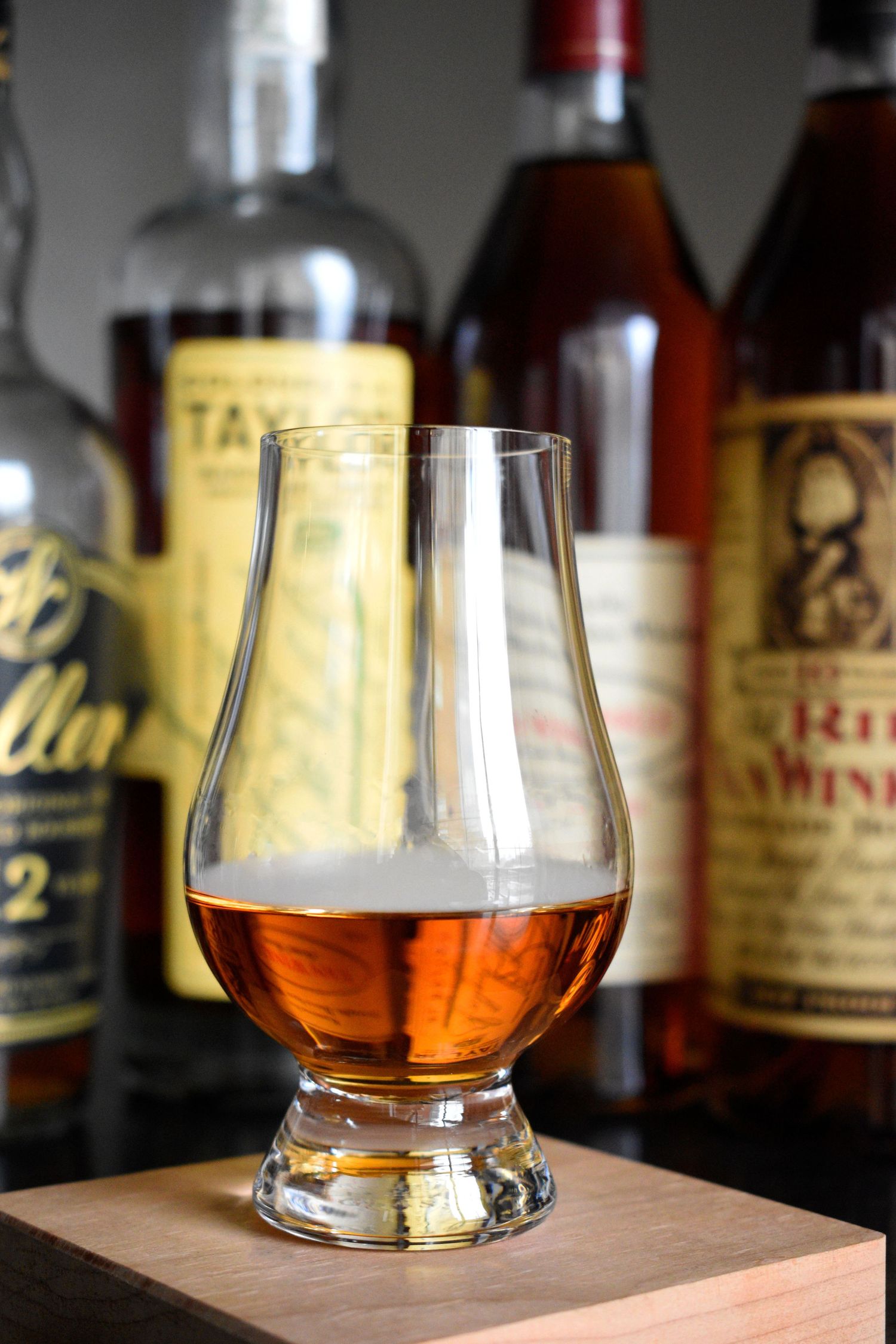 A Glencairn glass of amber bourbon whiskey on a wooden block surrounded by blurred bottles.