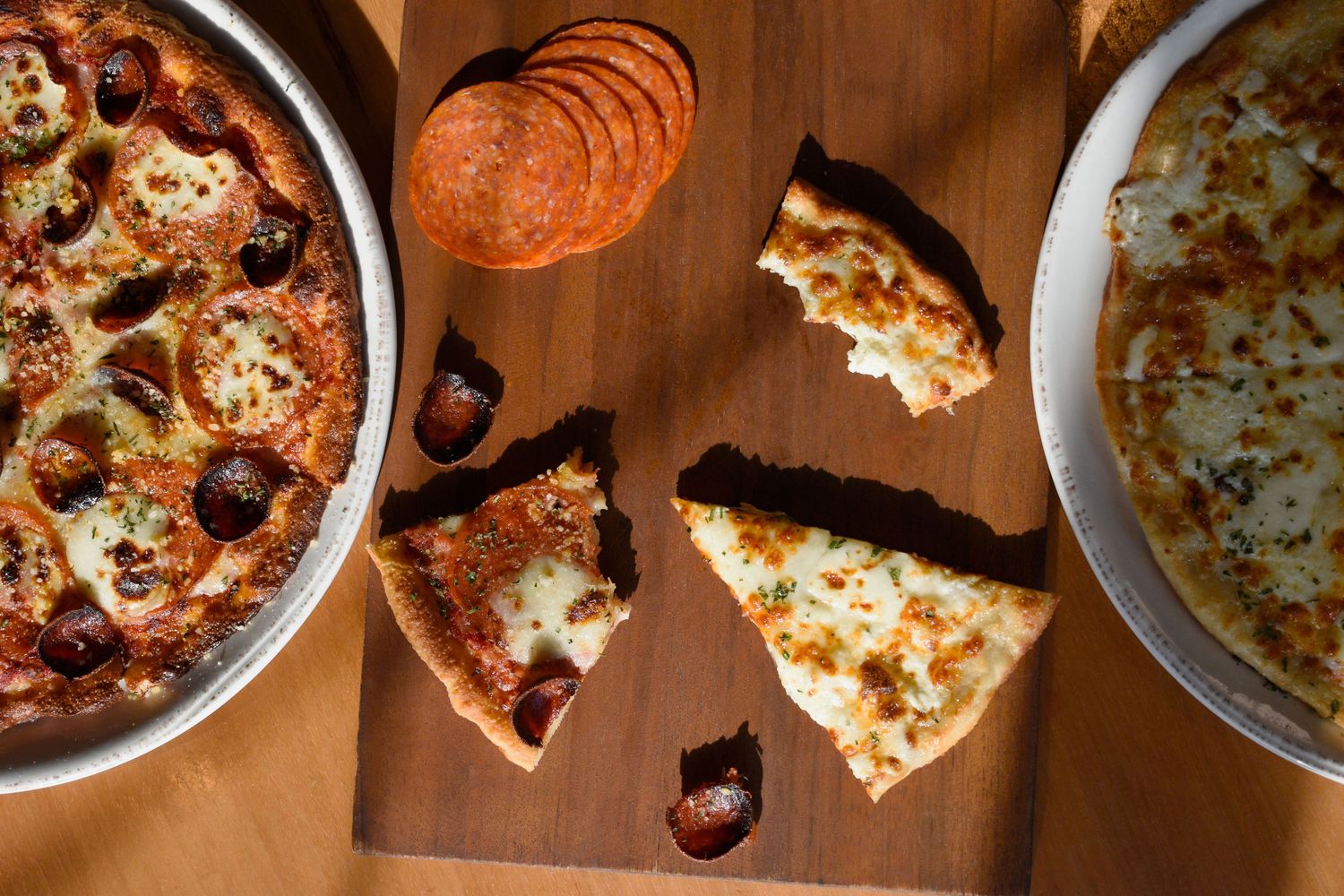 Sliced cheese and pepperoni pizza on a wooden board with two whole pizzas nearby.
