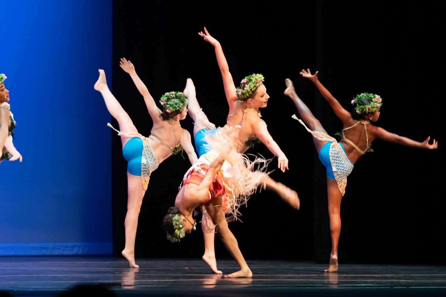 A group of dancers perform acrobatic moves in green and blue costumes on a bright stage.