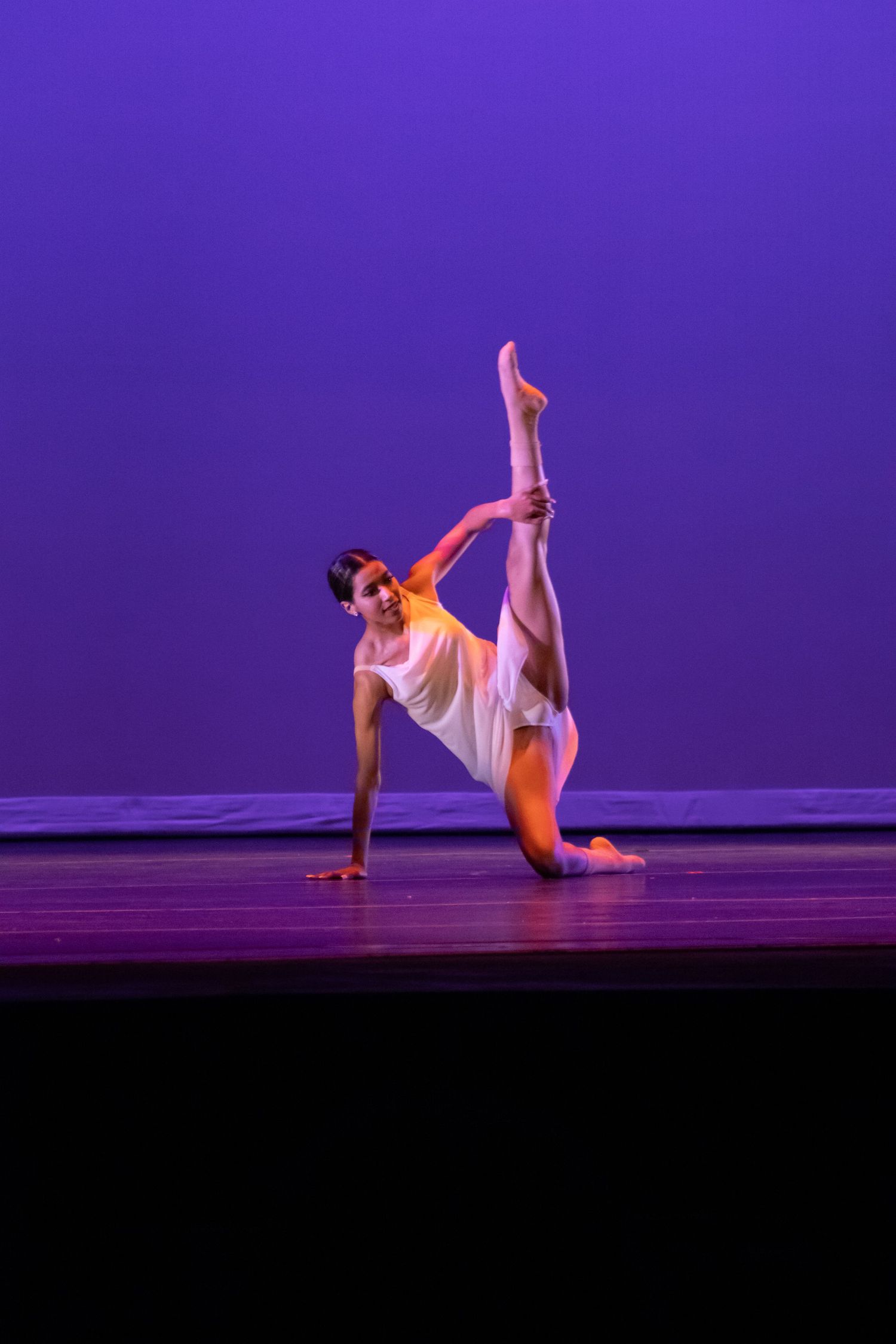 A flexible dancer performs a stunning leg extension on stage under purple lighting.