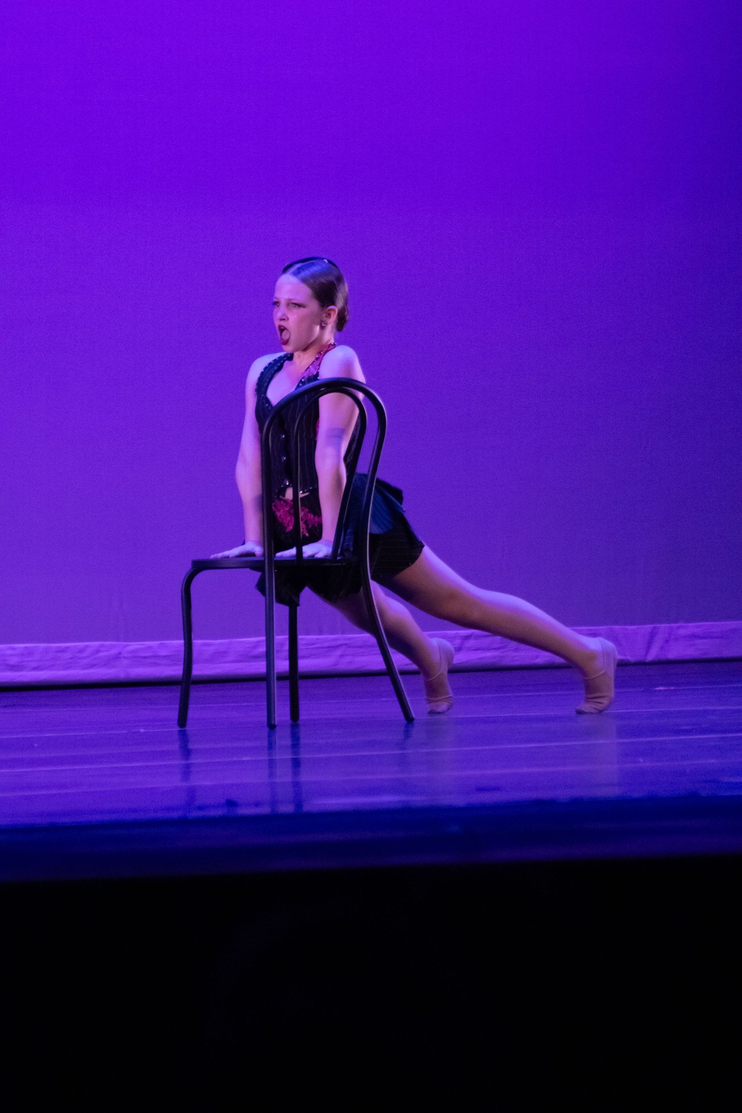 Female dancer in black leotard performs a lunge using a chair prop on a purple-lit stage.