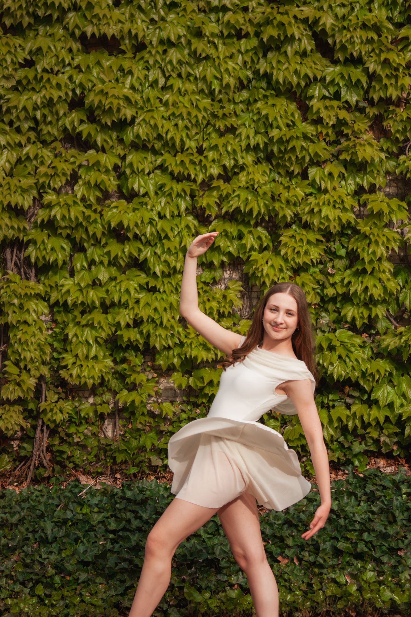 Young woman in white dress dancing gracefully in front of lush green ivy wall outdoors.