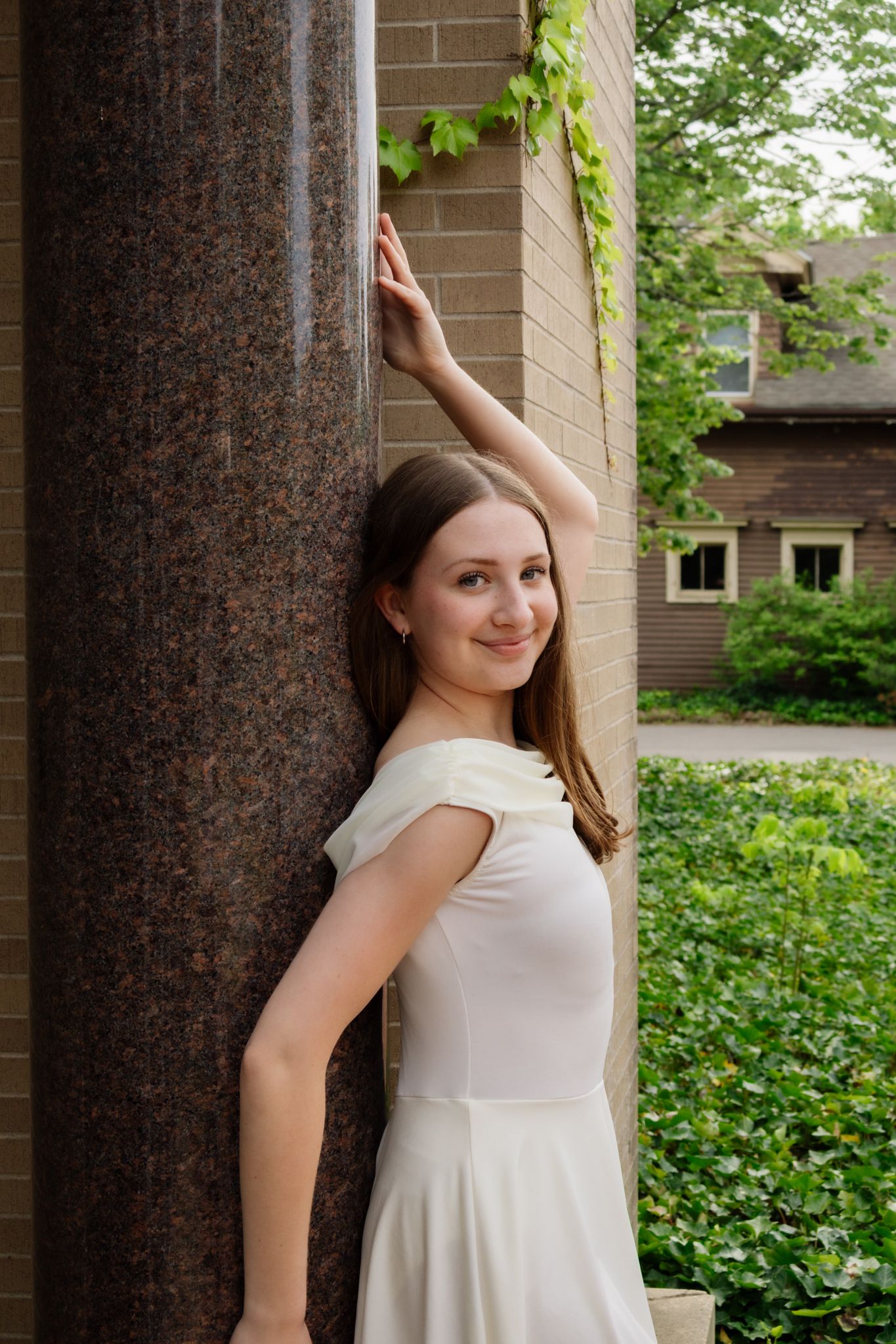 A young woman in a white dress smiles while posing beside a marble column with green ivy surroundings.